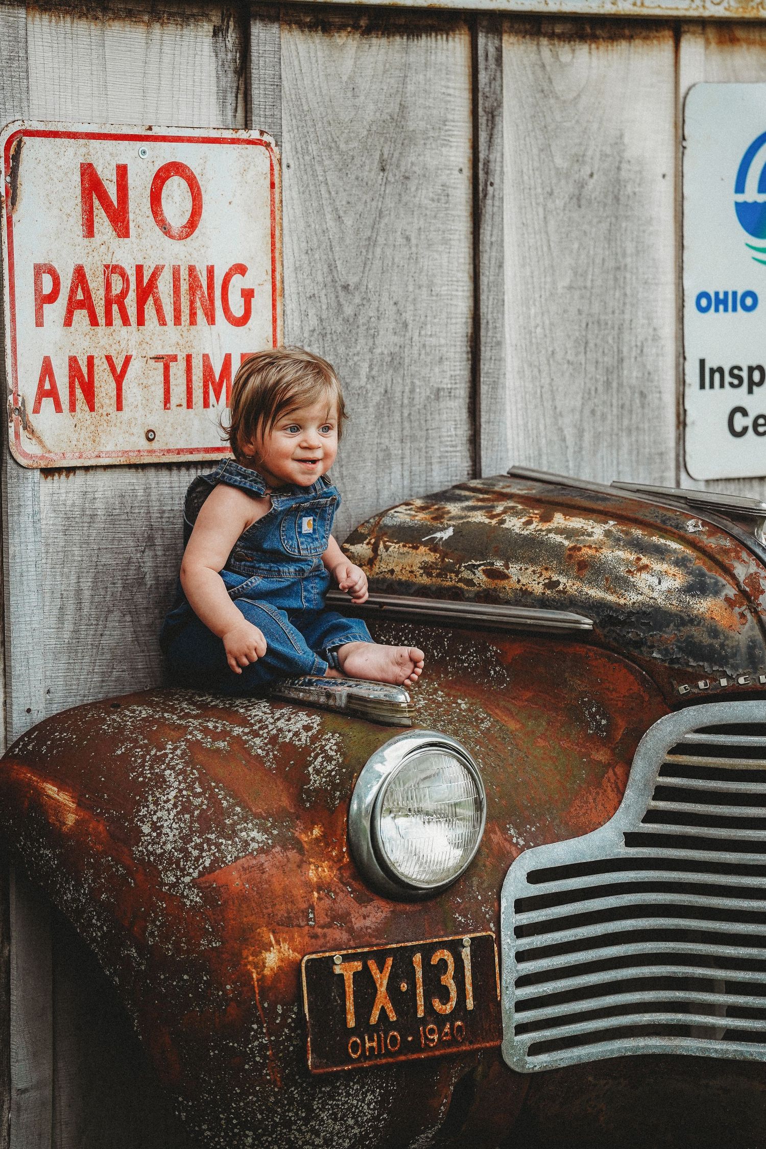 A rustic vintage car hood with peeling red paint and headlights is displayed near a No Parking sign.