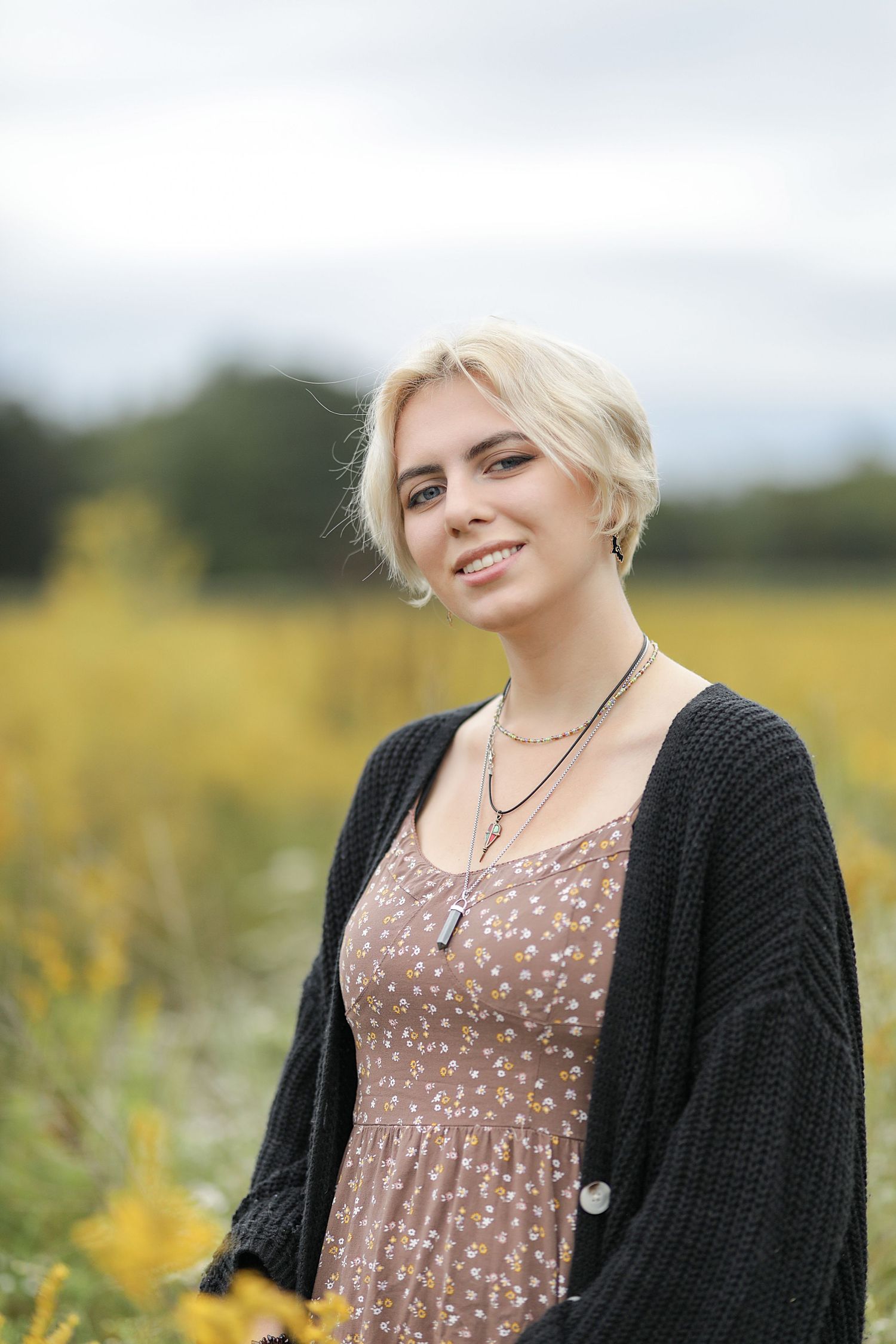 Person wearing black cardigan and polka dot dress stands in yellow wildflower field on sunny day outdoors.