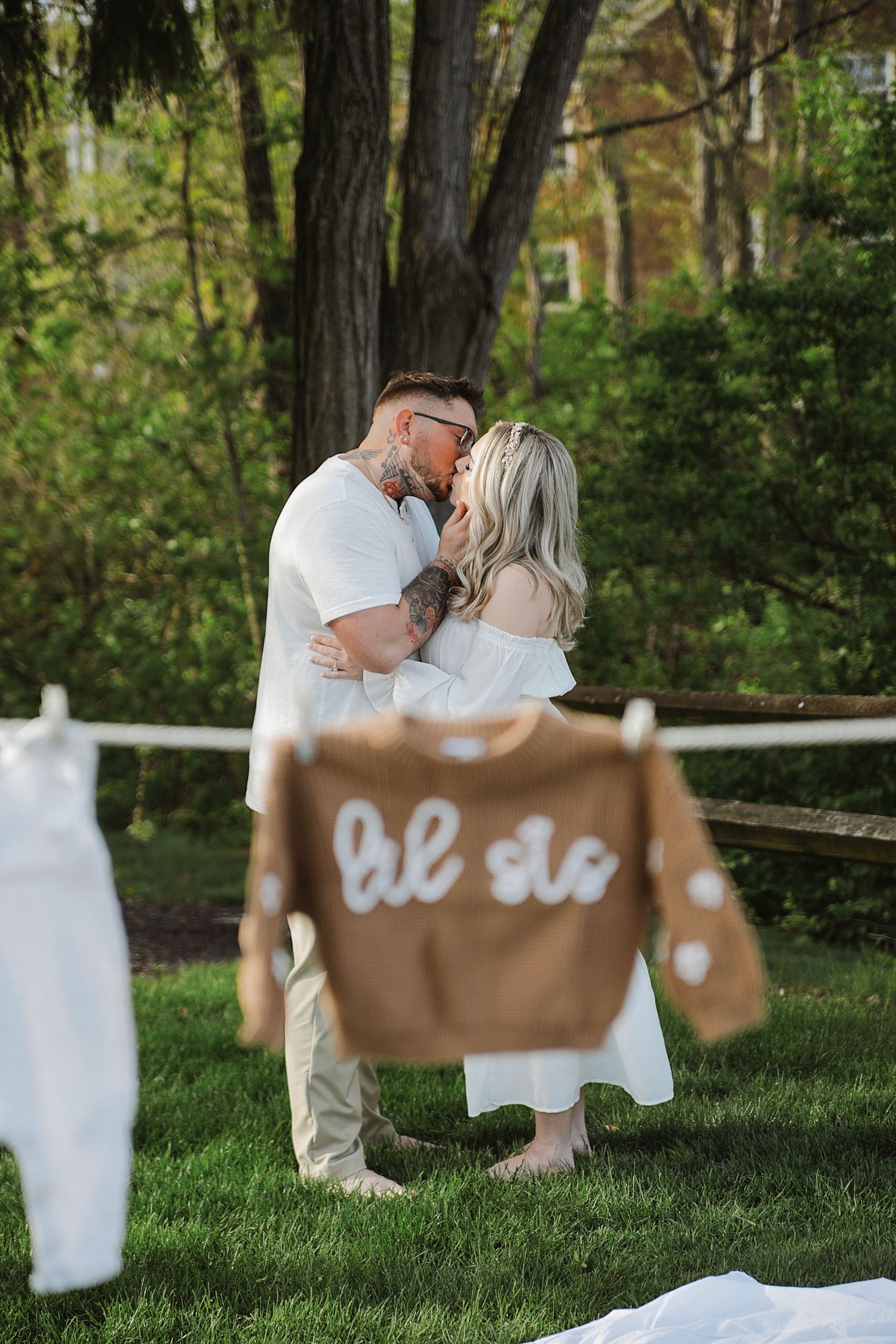 Romantic outdoor engagement photo with rustic 'be so' sign hanging on clothesline while couple embraces in garden setting.