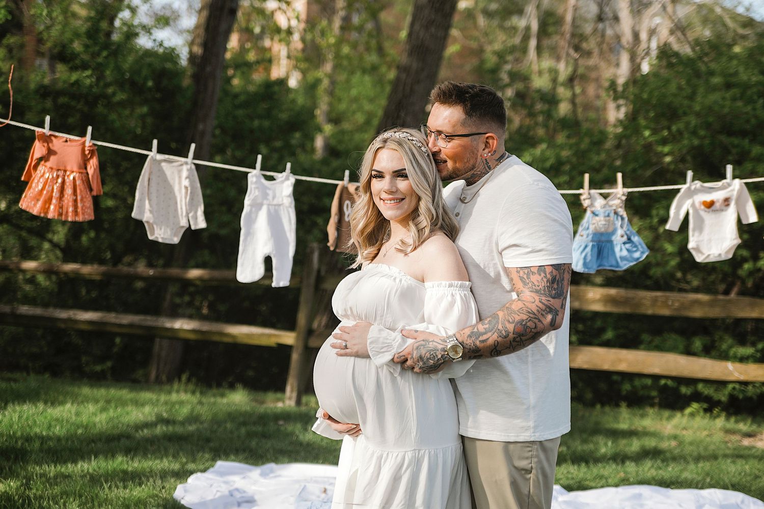 Expecting parents pose in a garden with baby clothes hanging on clothesline during maternity photoshoot.