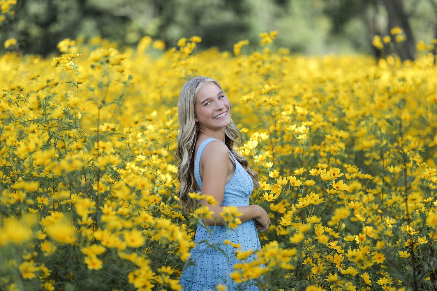 A joyful figure wearing a light blue top stands amidst a vibrant field of blooming yellow wildflowers on a sunny day.