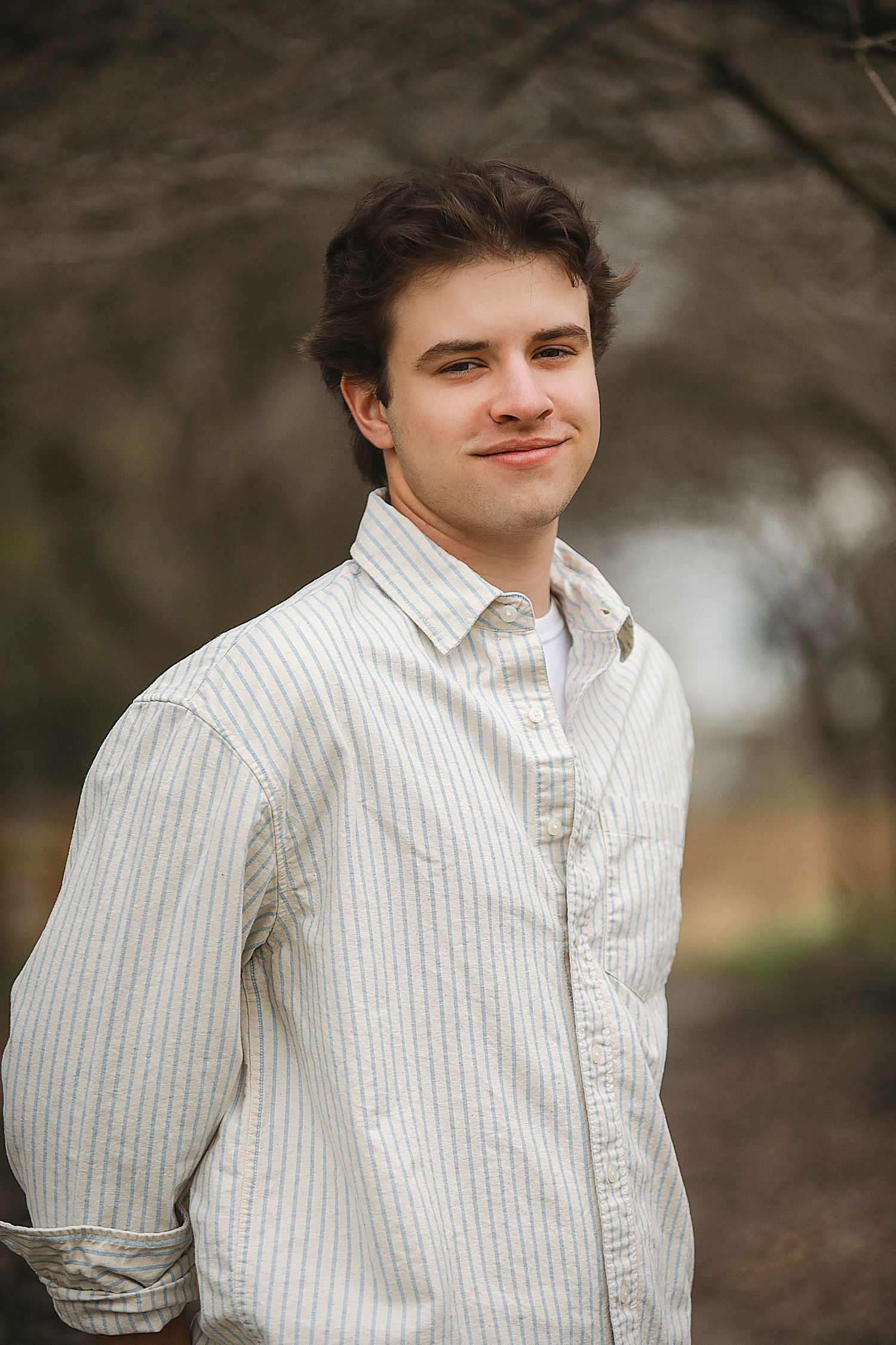 Person in a white pinstriped button-down shirt poses outdoors against a blurred natural background.