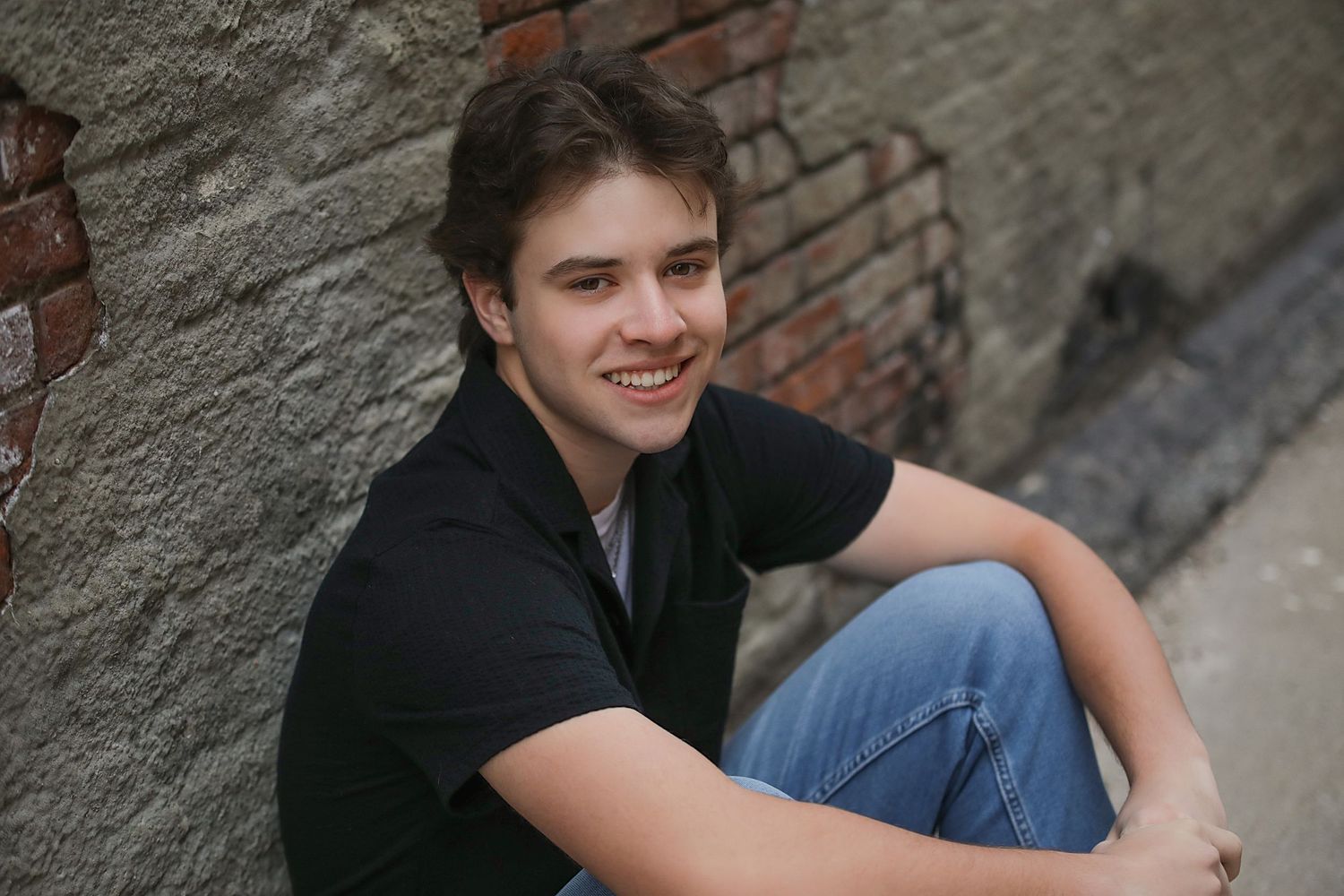 Young person in black polo shirt sitting casually against concrete wall with brick detail.