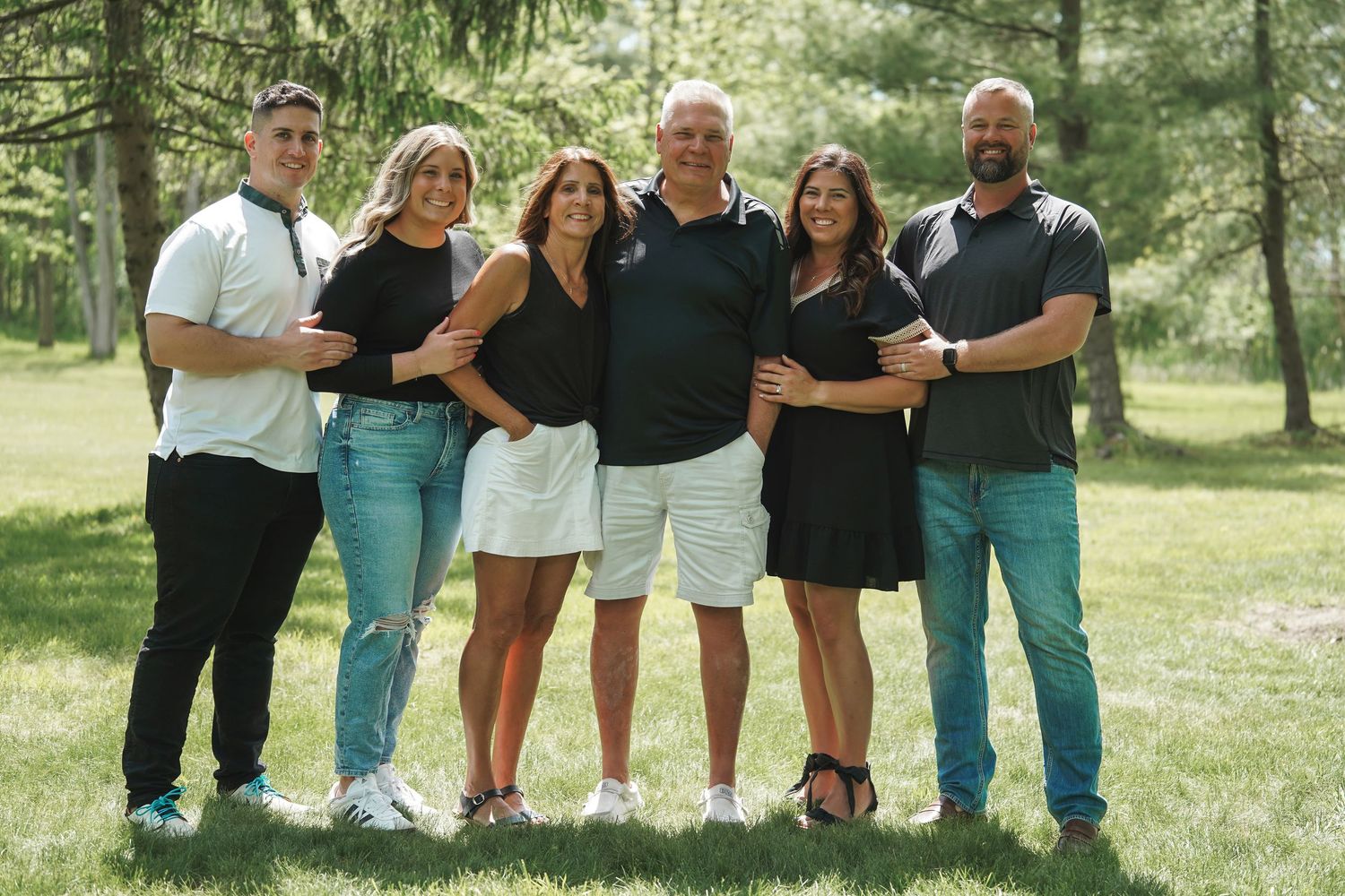 A group of six people in casual attire pose together outdoors on a sunny day with green trees in the background.