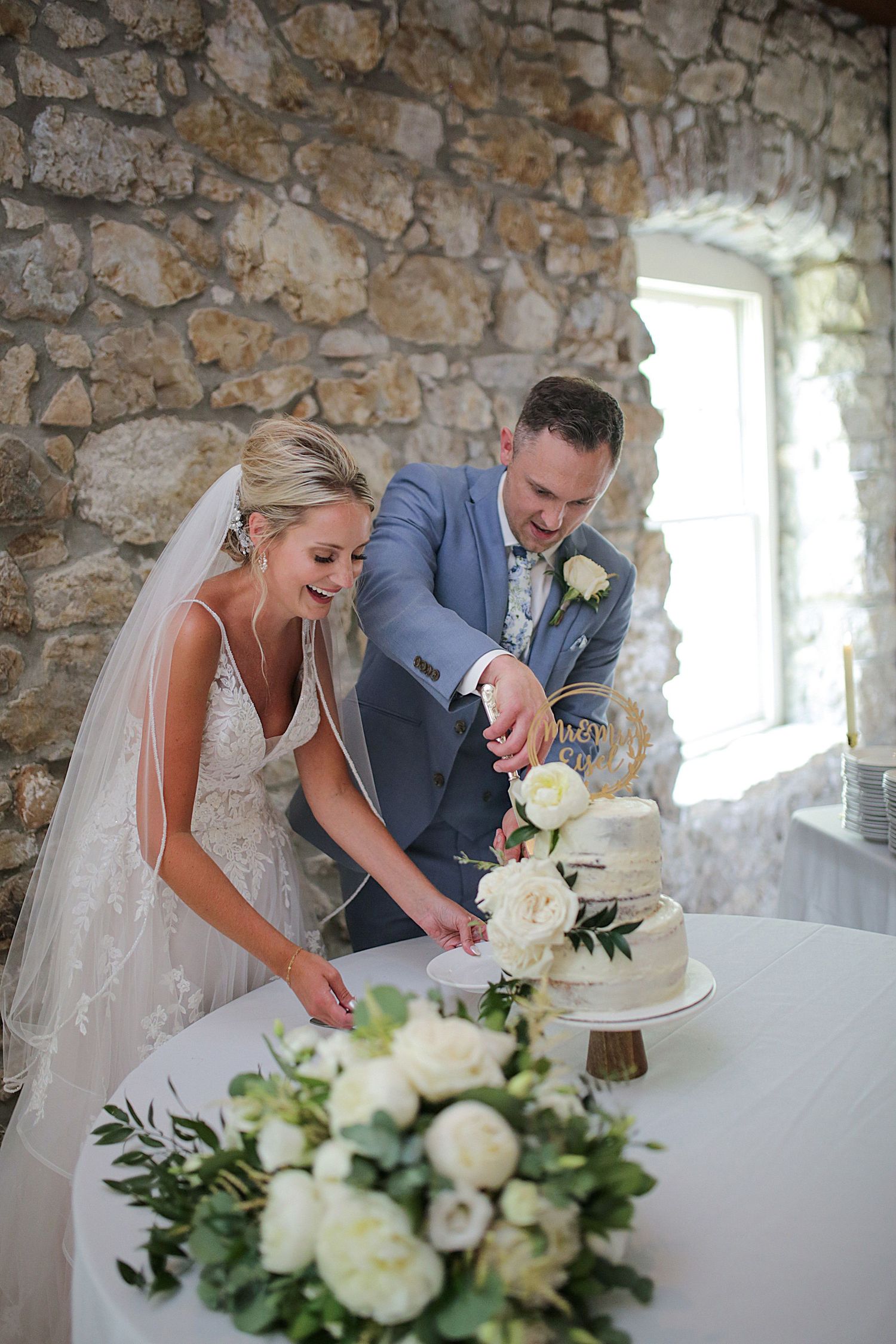 Newlyweds cut their elegant white wedding cake together against a rustic stone wall.