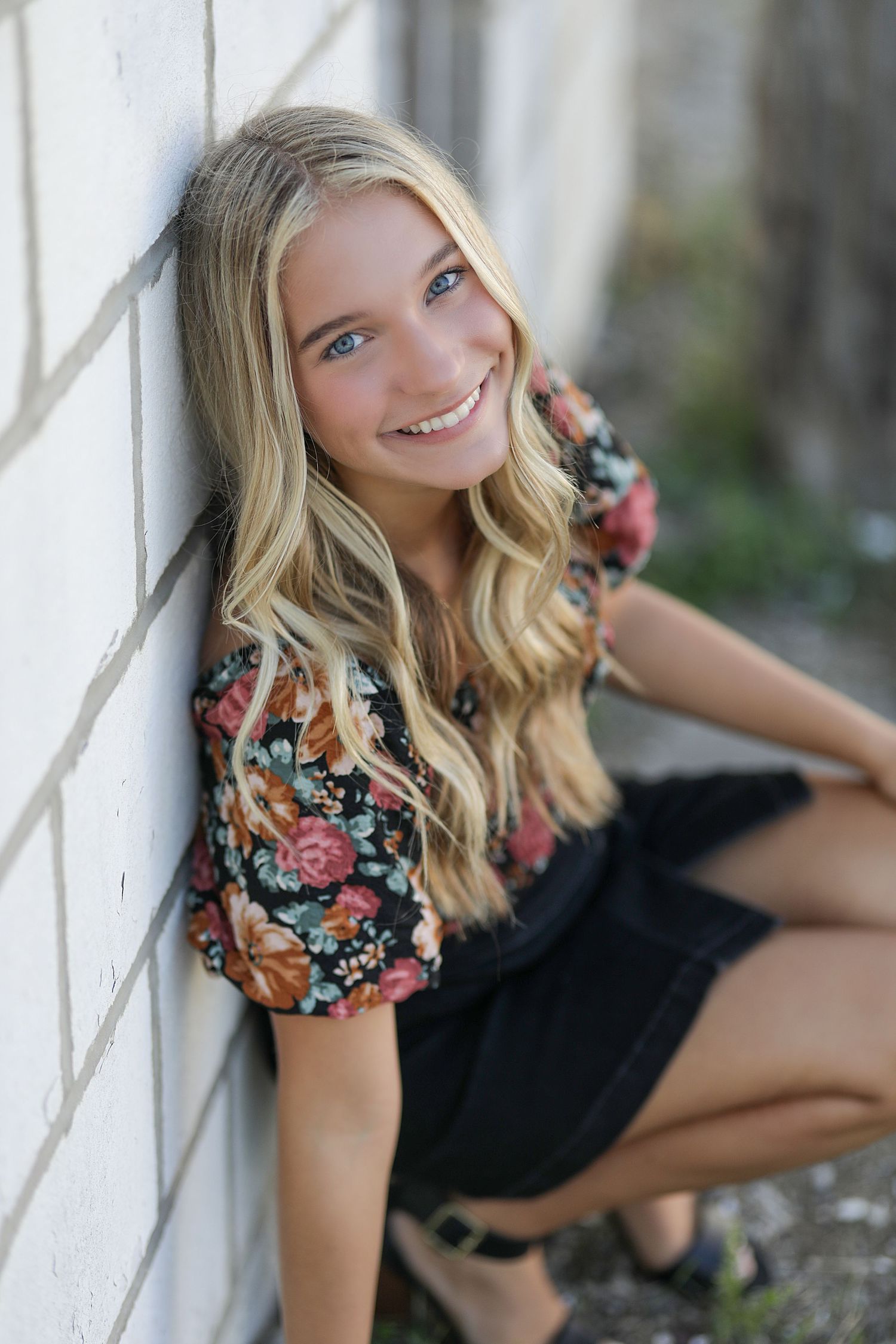 Outdoor portrait of someone in a floral top and black skirt sitting against a white wall with wavy blonde hair.