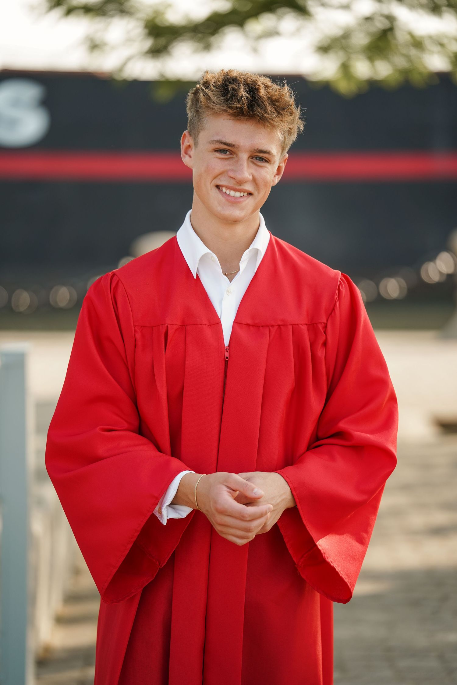 Graduate wearing bright red graduation gown and white dress shirt poses outdoors in sunlight.