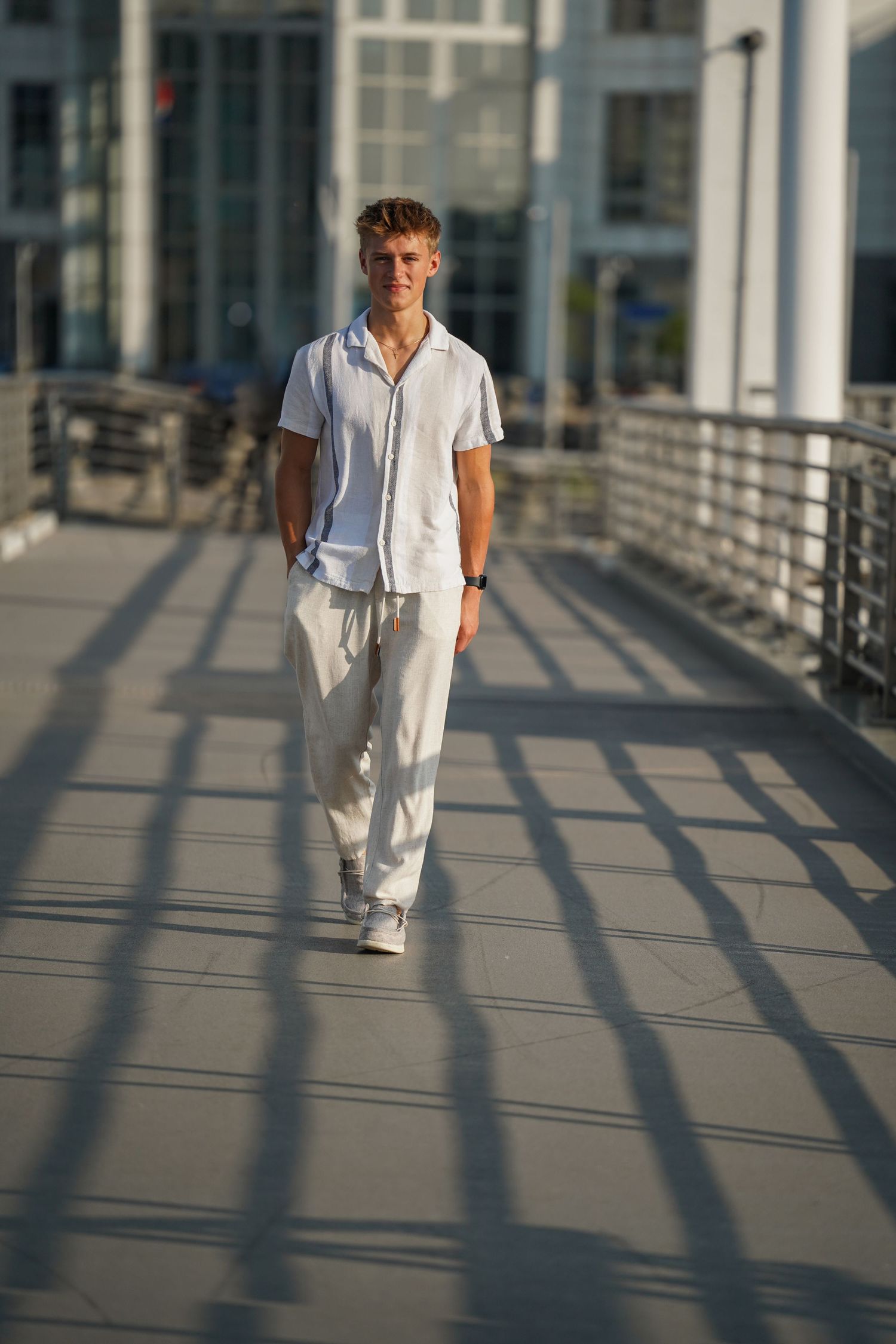 A person in all-white summer attire walks on a modern walkway during golden hour casting long shadows.