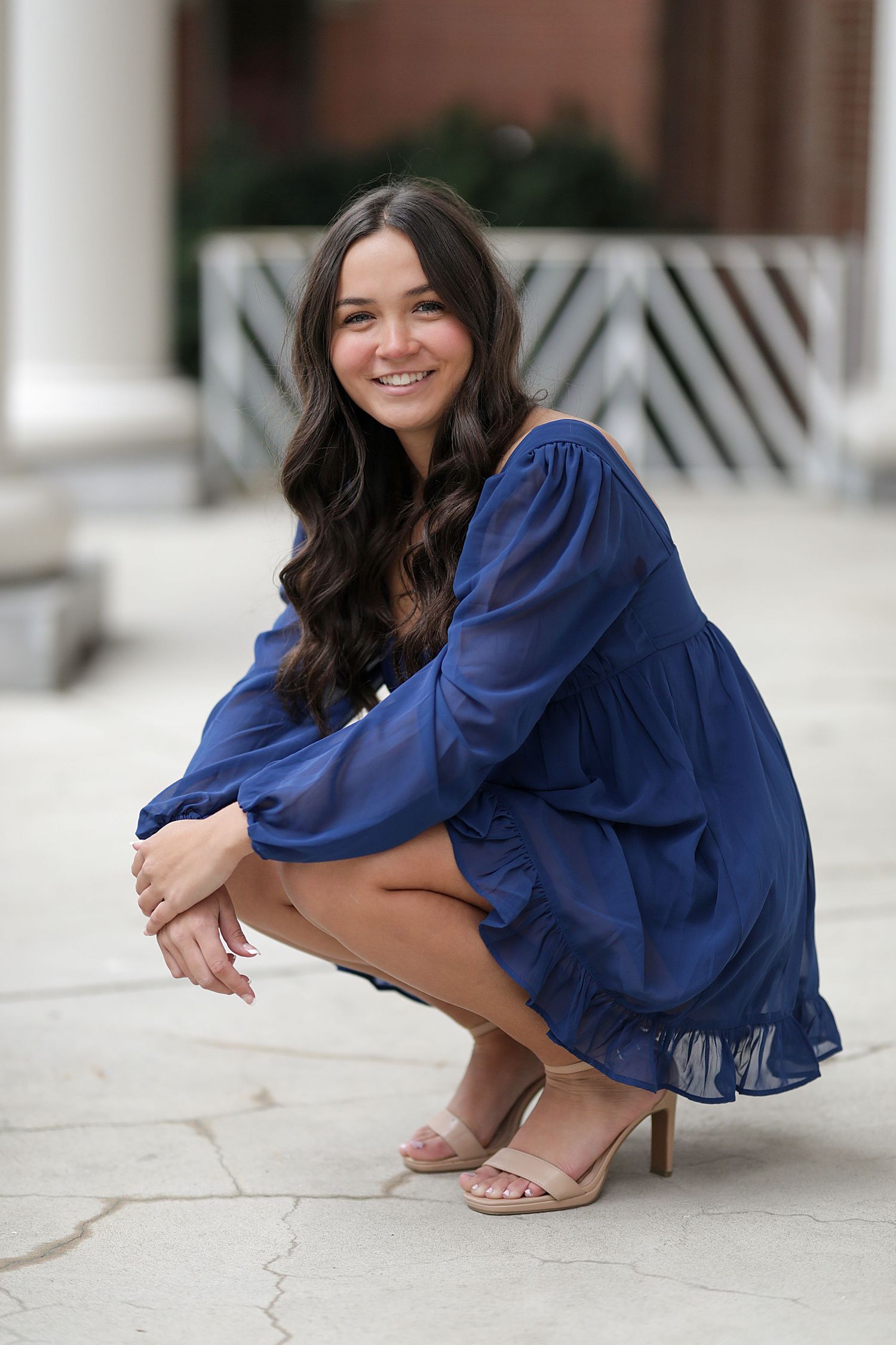 Woman in flowing navy blue dress and beige sandals gracefully poses on concrete surface in outdoor photo series.