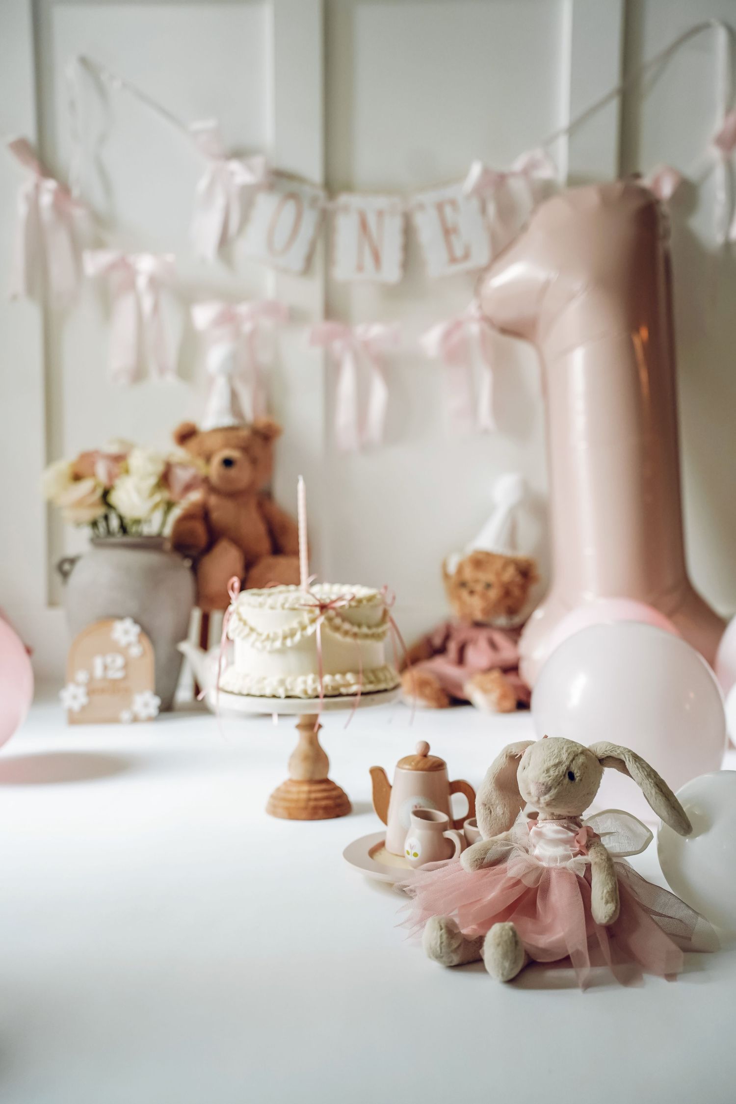 First birthday party decorations with pink balloon, teddy bears, small cake on stand, and bunting banner in soft pastel colors.