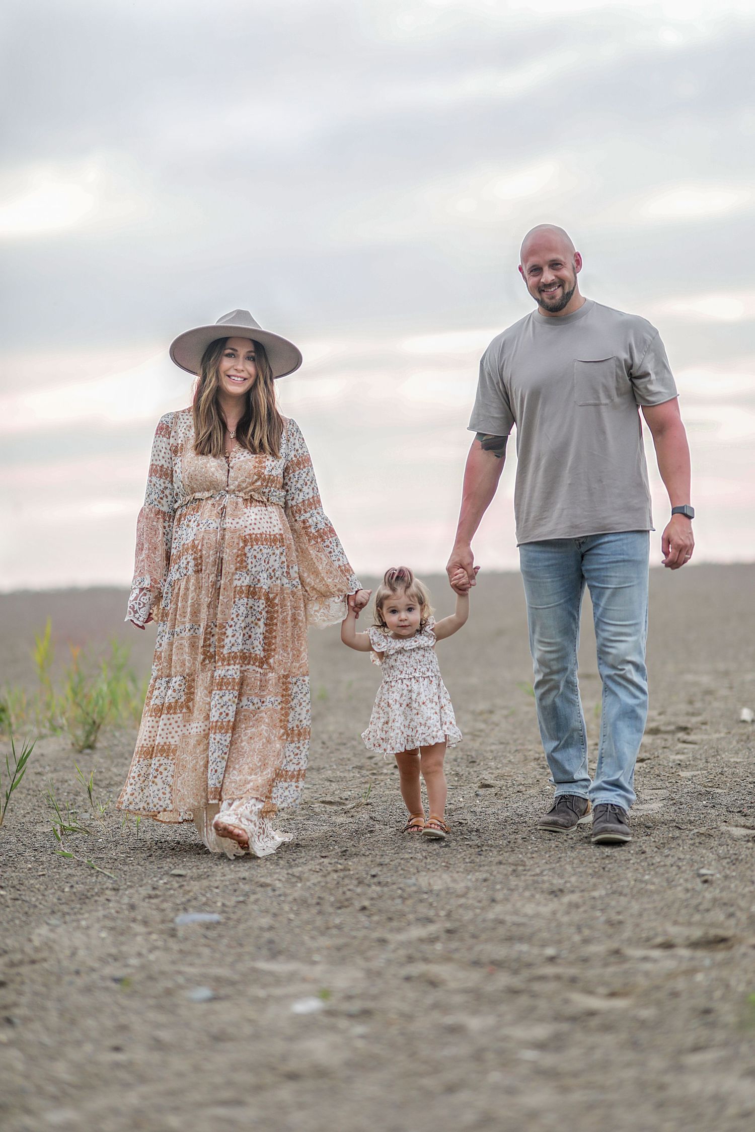 A family walks together on a dirt path at sunset with the mother wearing a flowy dress and wide-brimmed hat.