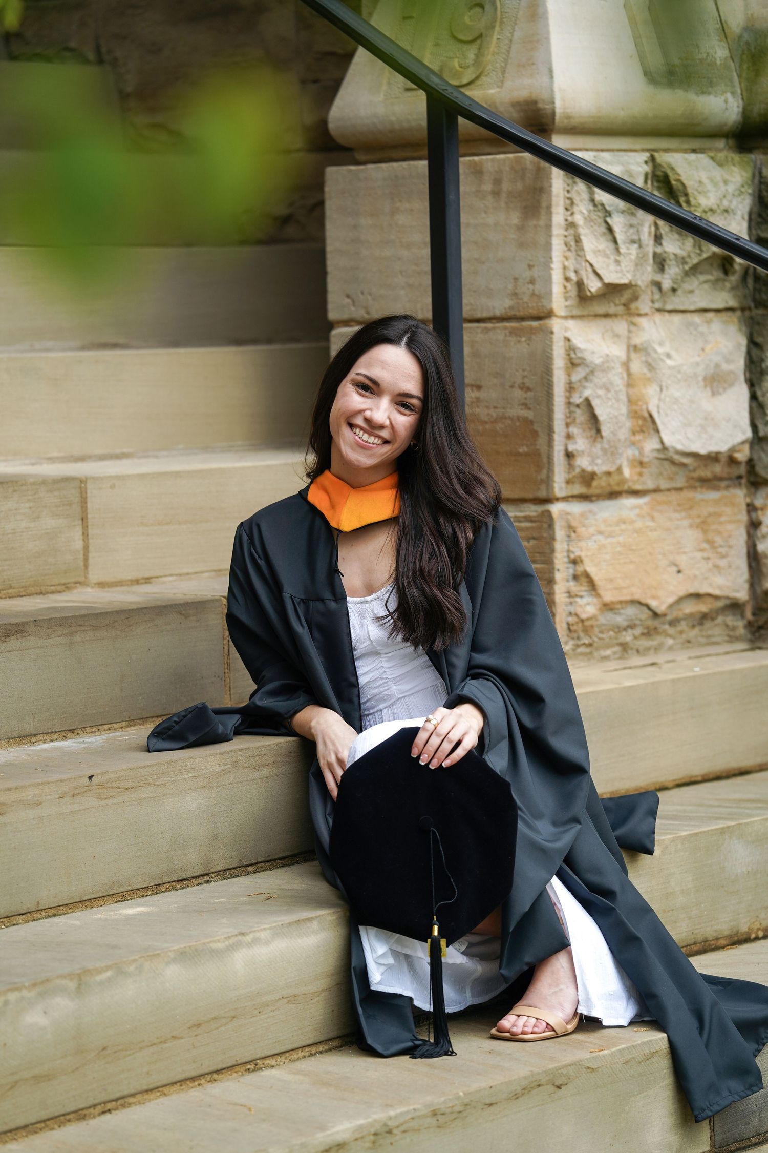 Graduate in black and white attire sits casually on outdoor stone steps at a university campus.