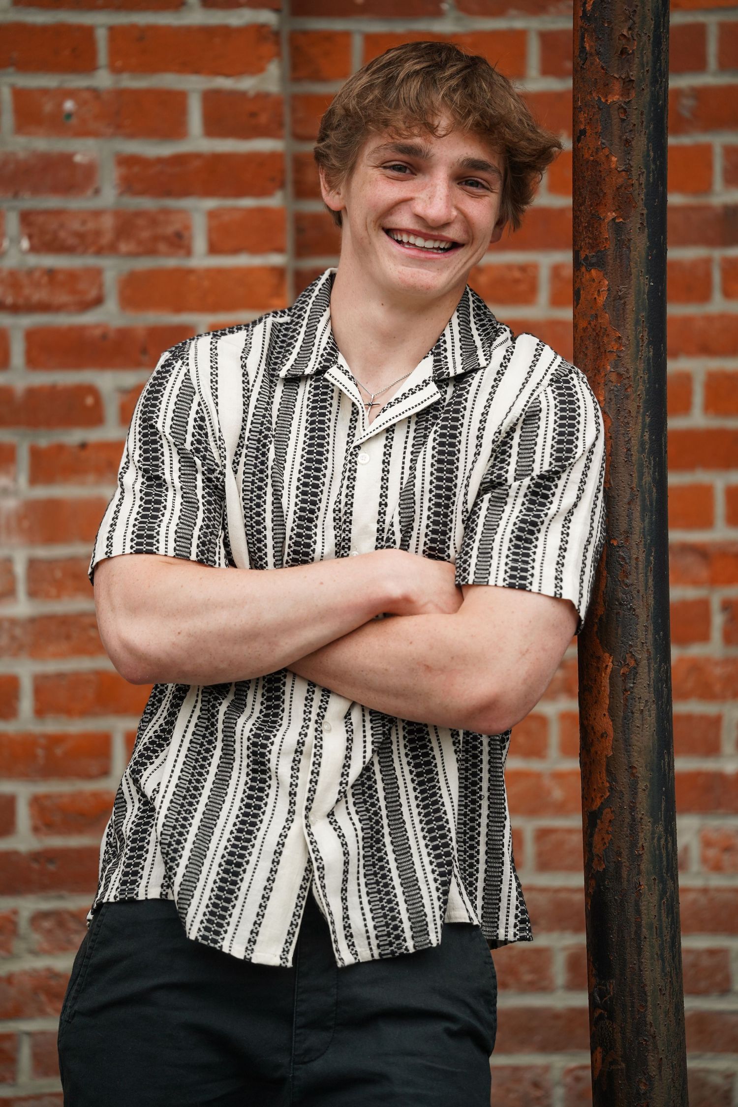 A person in a black and white striped shirt poses with crossed arms against a rustic brick wall.