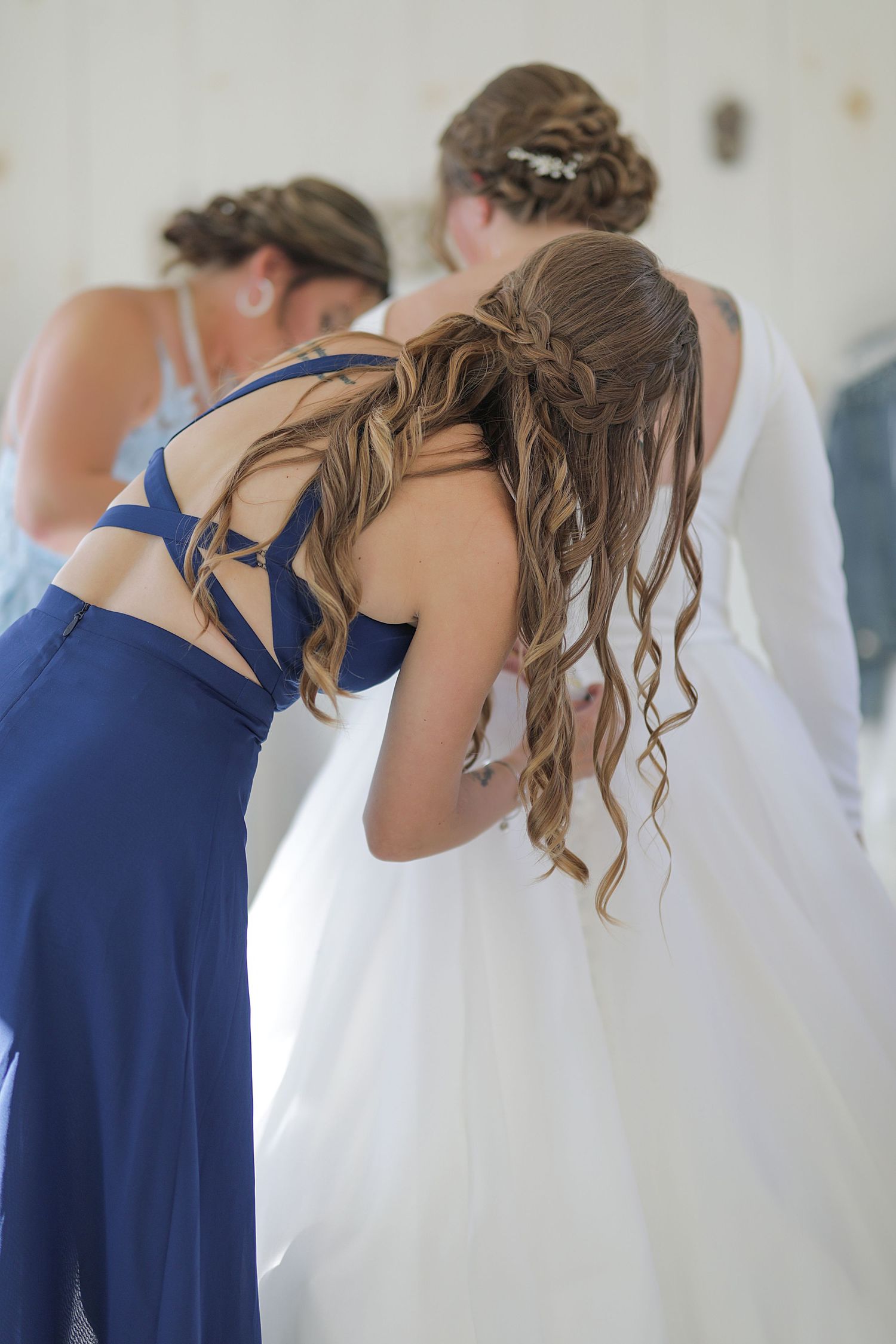 Bridesmaids in navy blue dresses and a bride in white gather together showing intricate hairstyles from behind.