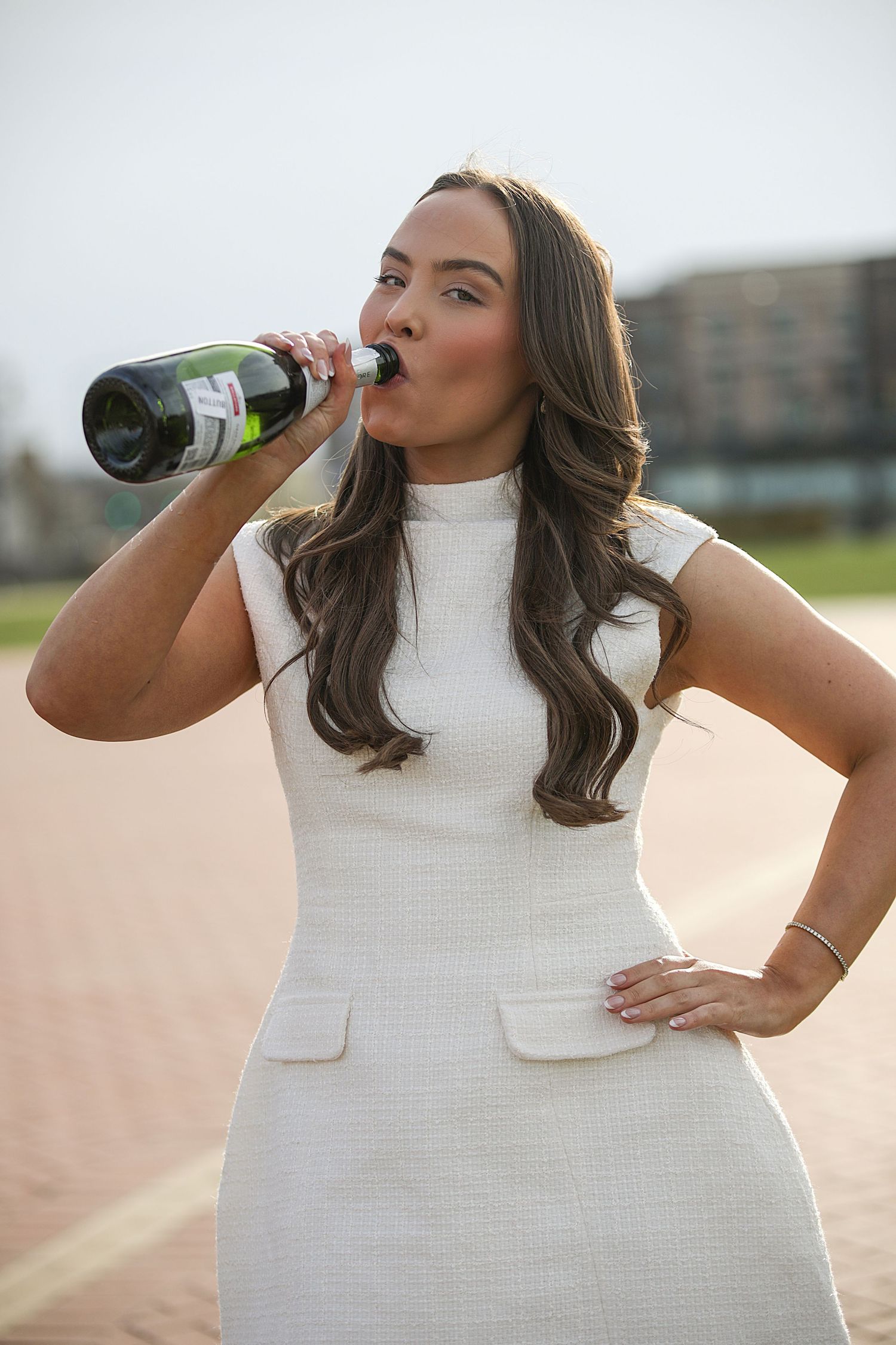 Woman in white sleeveless top takes a sip from a dark glass bottle during outdoor photoshoot.