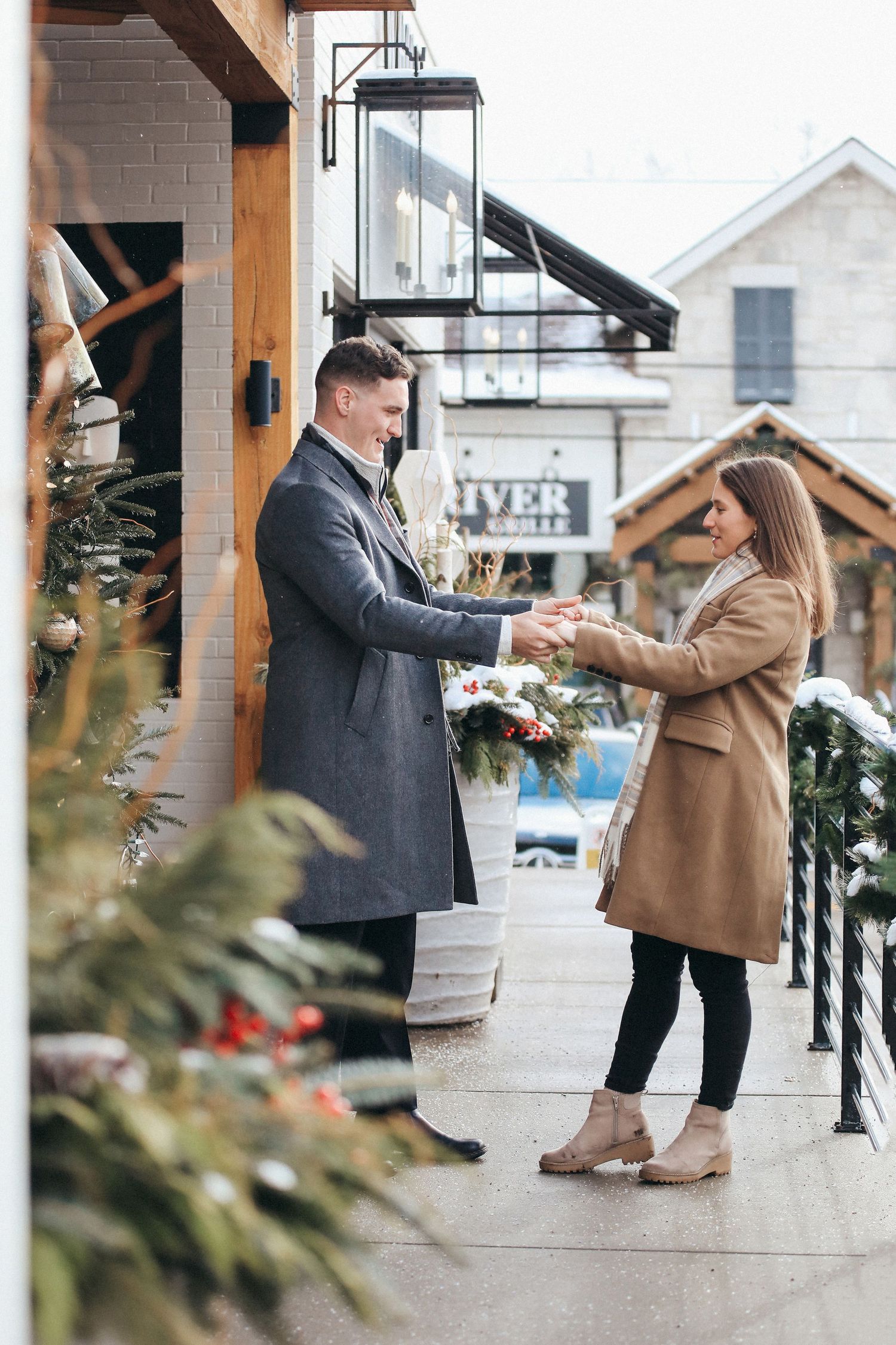 Couple in winter coats holding hands on snowy porch with holiday decorations.