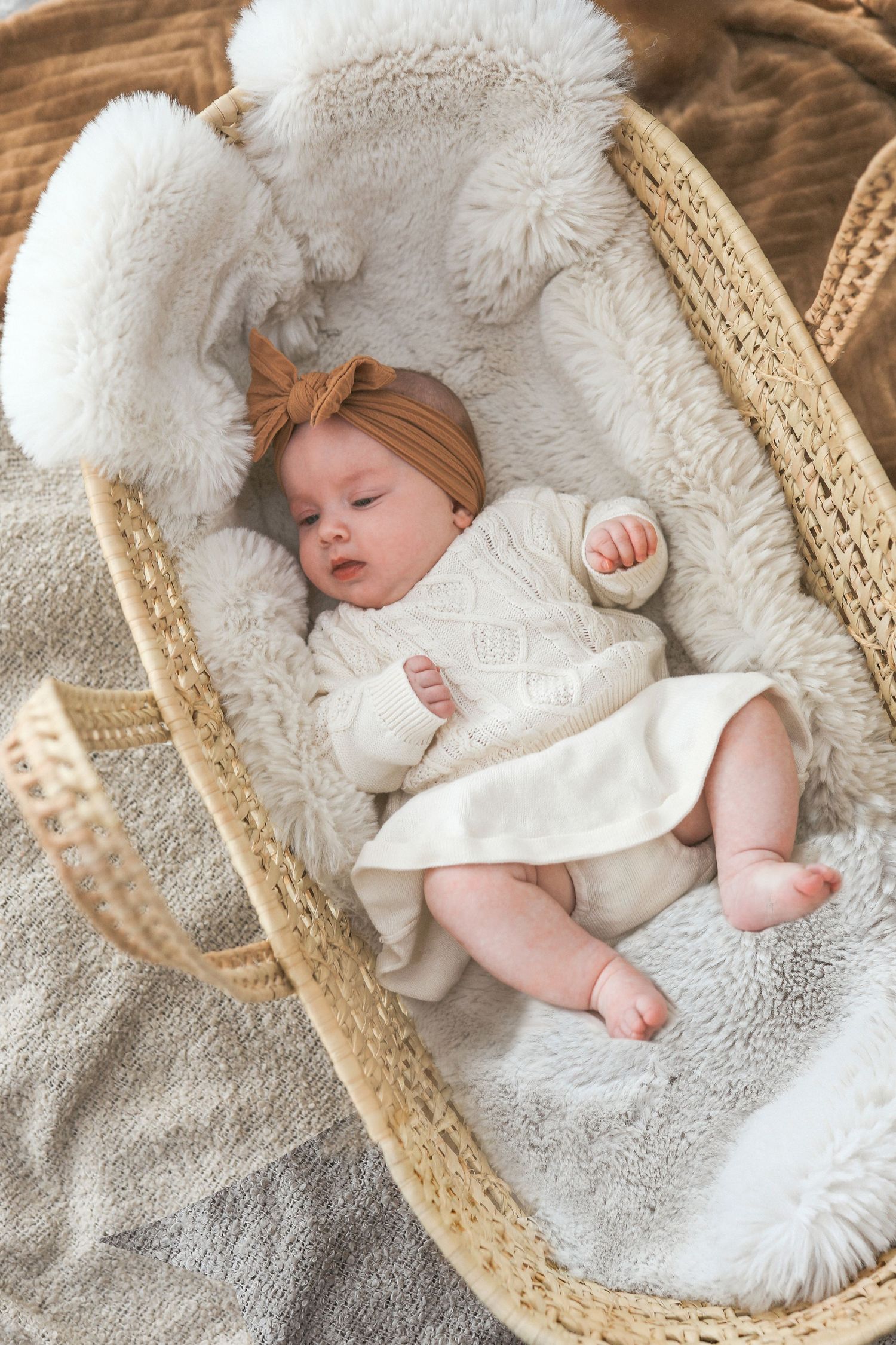 A sleeping baby rests peacefully in a woven bassinet lined with soft white fur bedding.