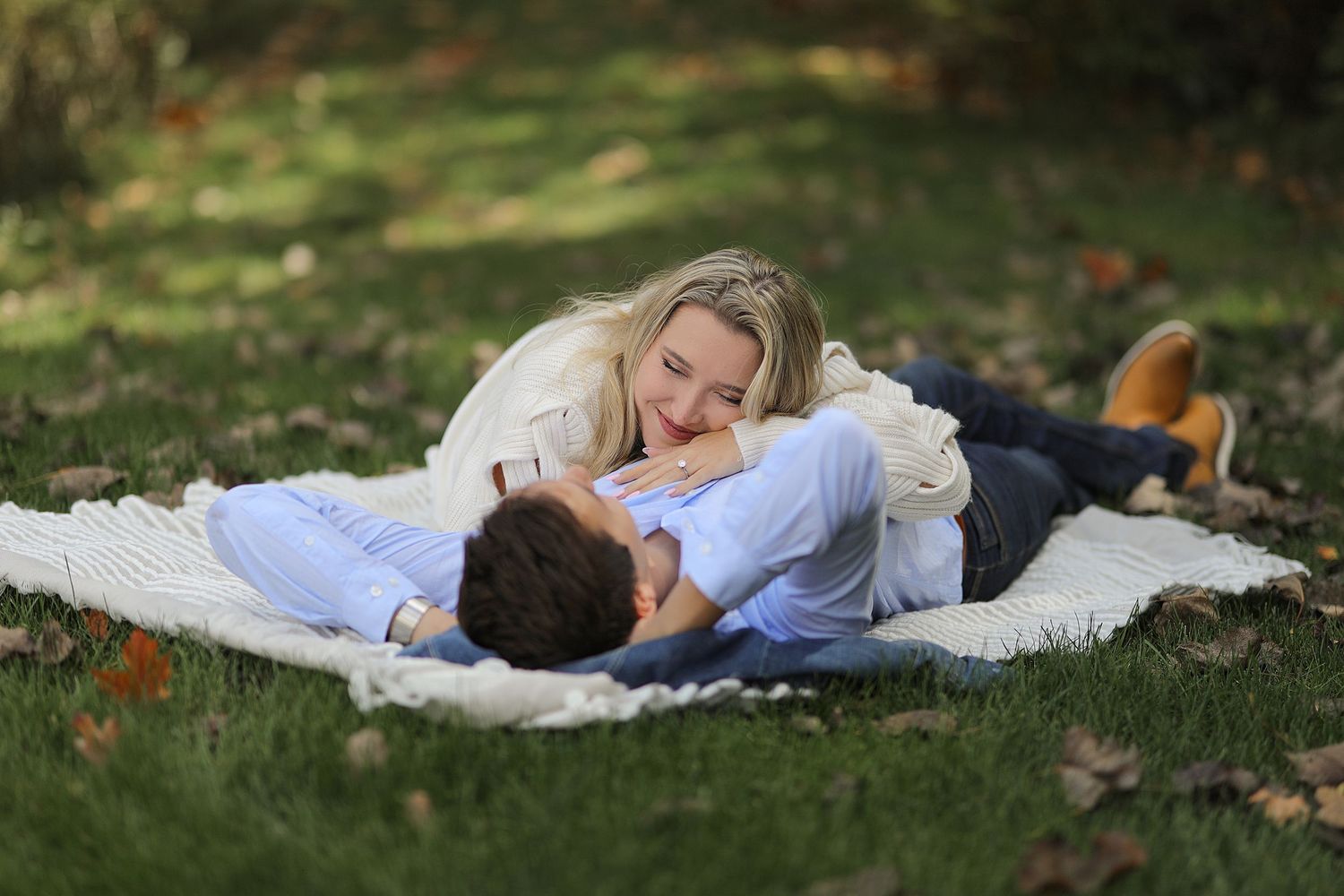 Two people share a romantic moment while laying on a white blanket in grass surrounded by fallen autumn leaves.