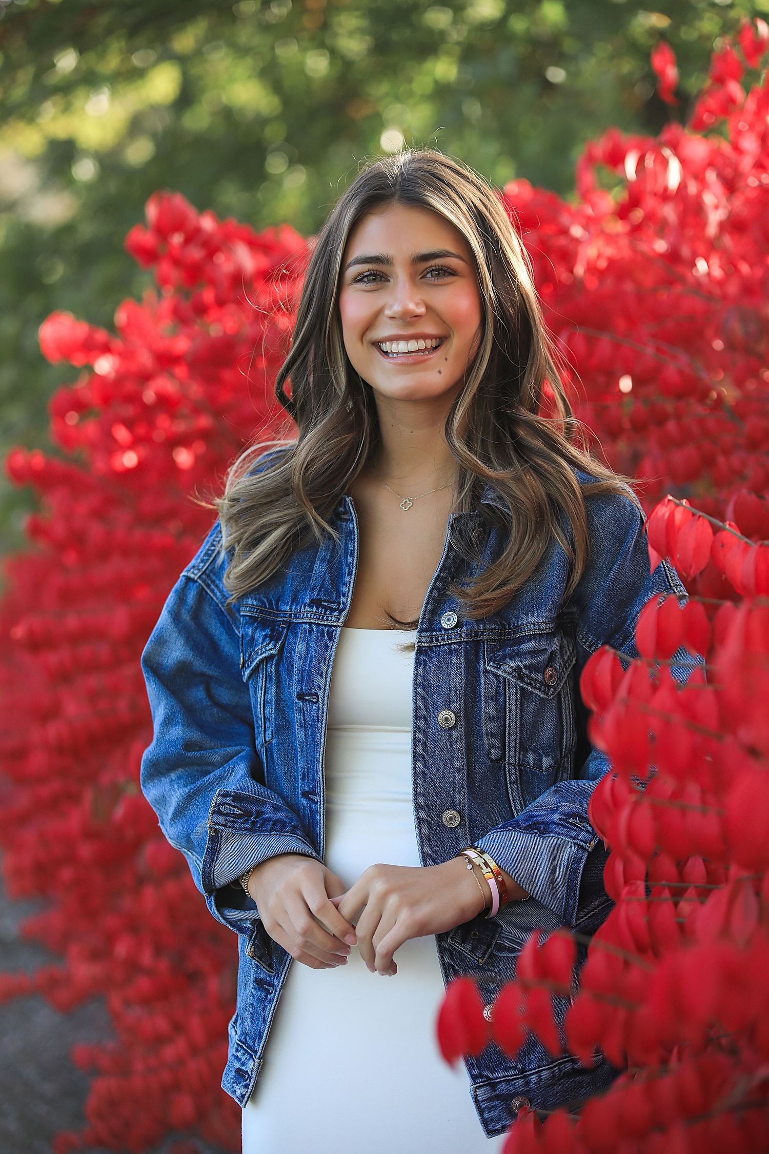 Fashion portrait outdoors showing denim jacket styled with white top against vibrant red autumn foliage background.
