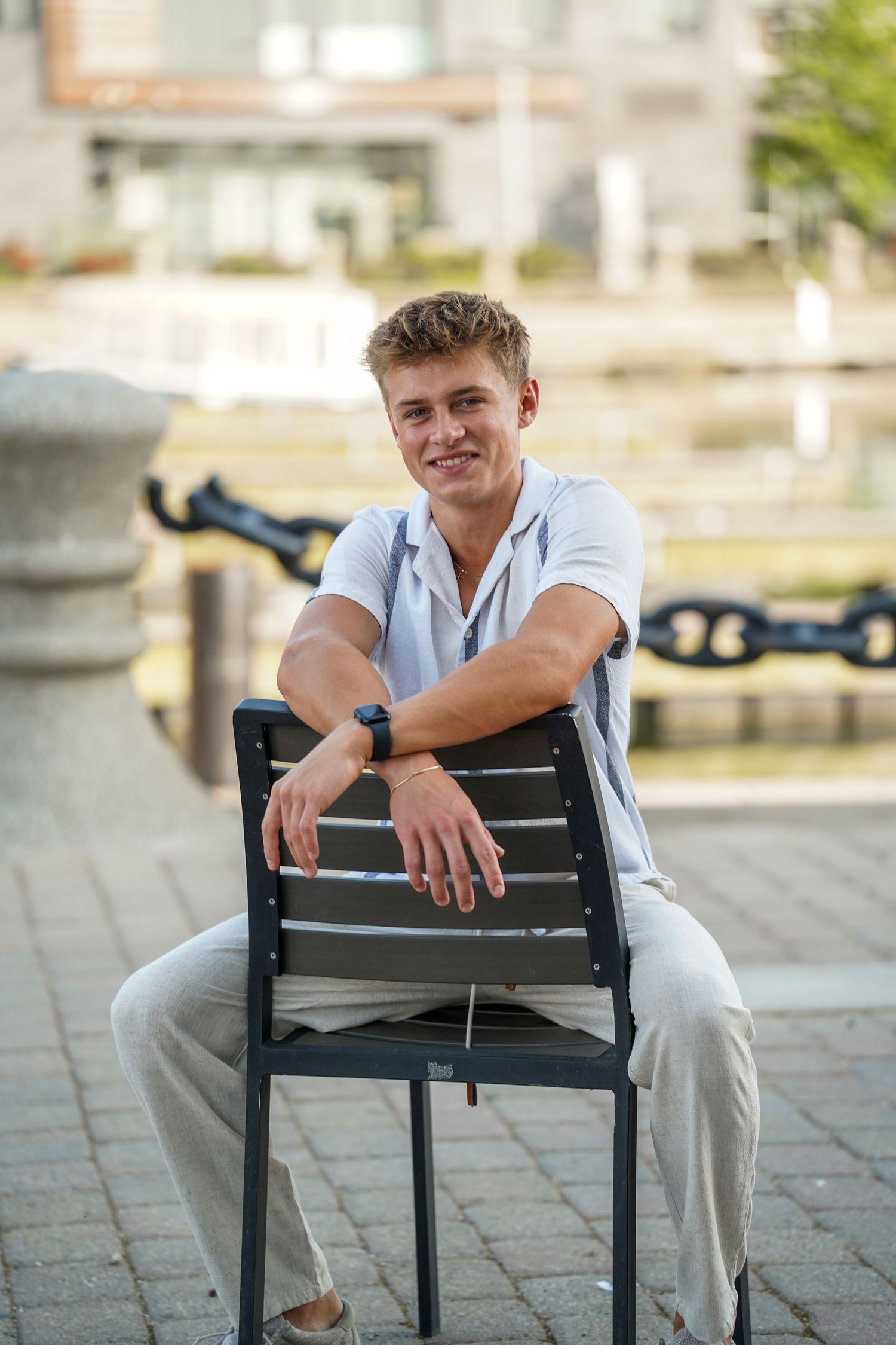 A person sits casually on a black chair outdoors wearing a light colored shirt and pants in a casual outdoor setting.