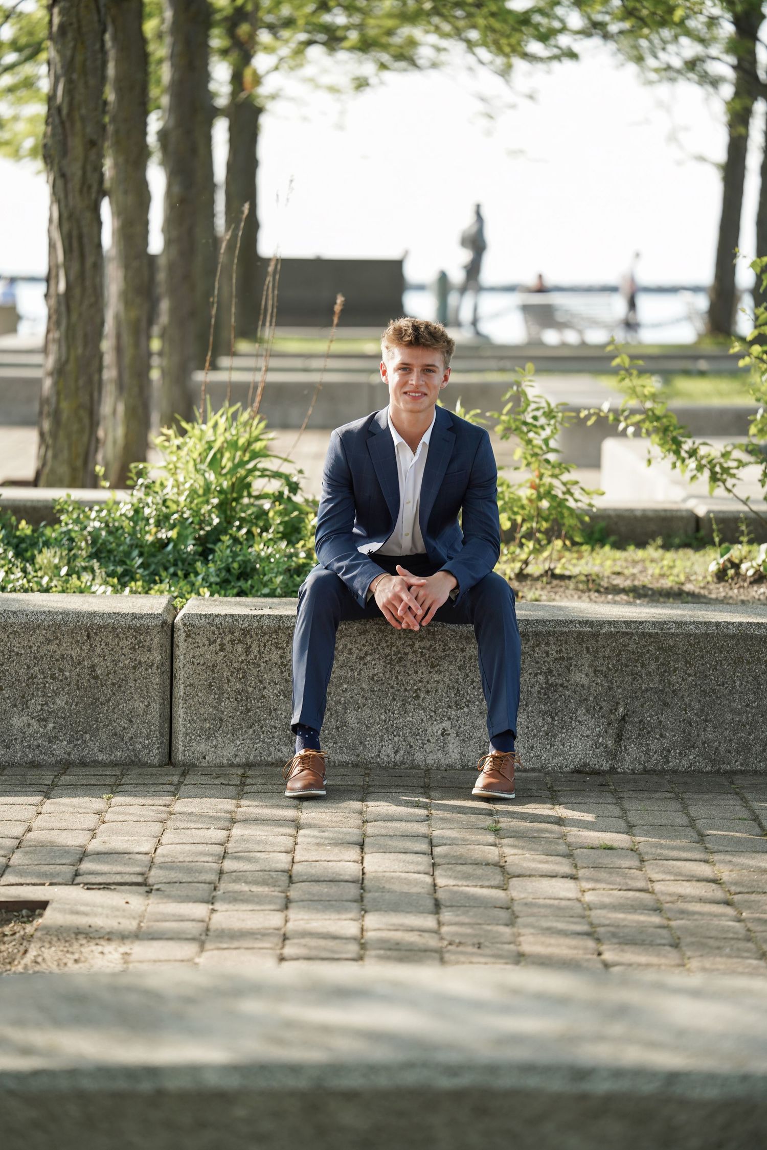 Young professional in navy suit relaxes on stone wall in park setting with trees and water views on sunny day.