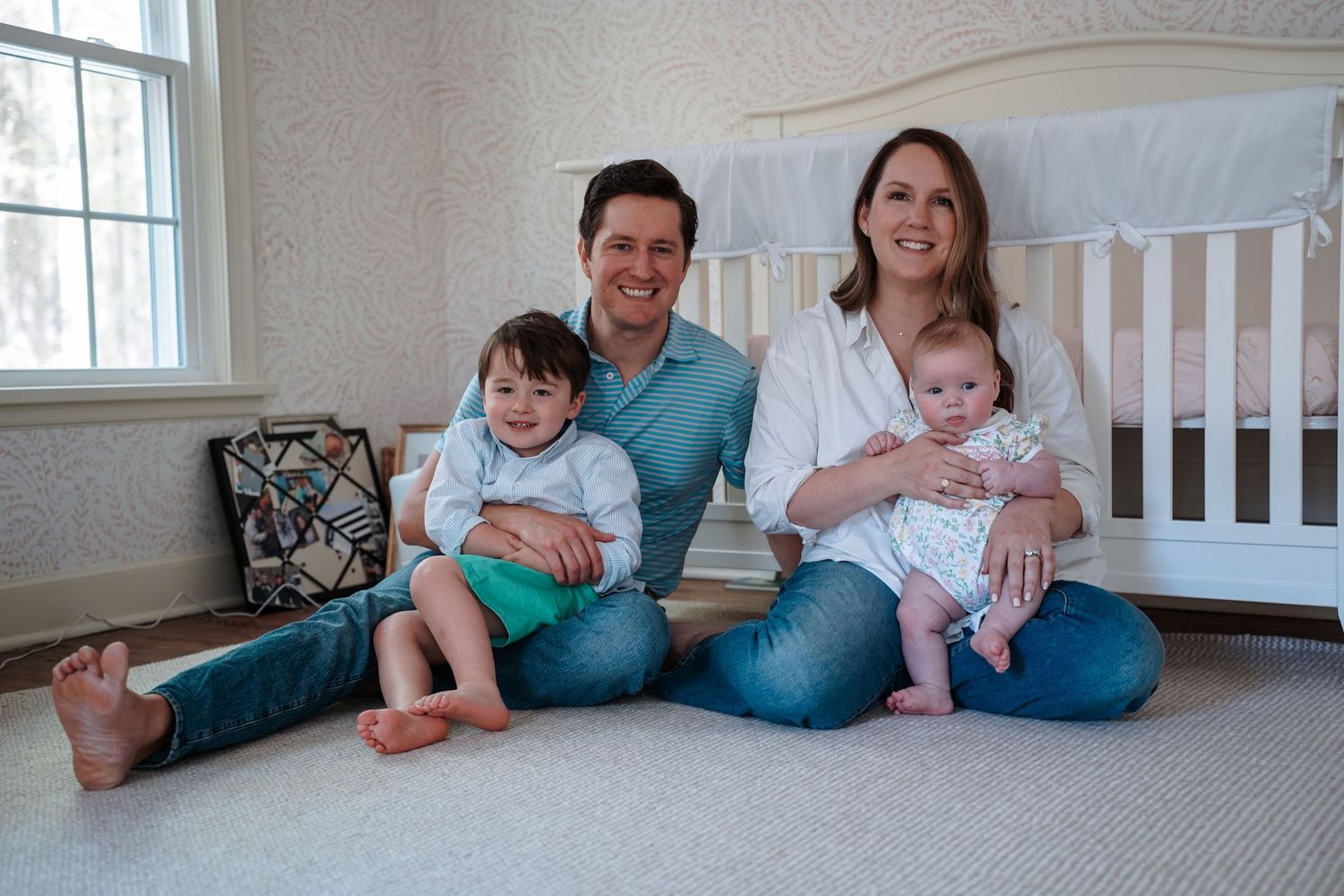 Family sitting together on carpet in nursery room with white crib and floral wallpaper in background.
