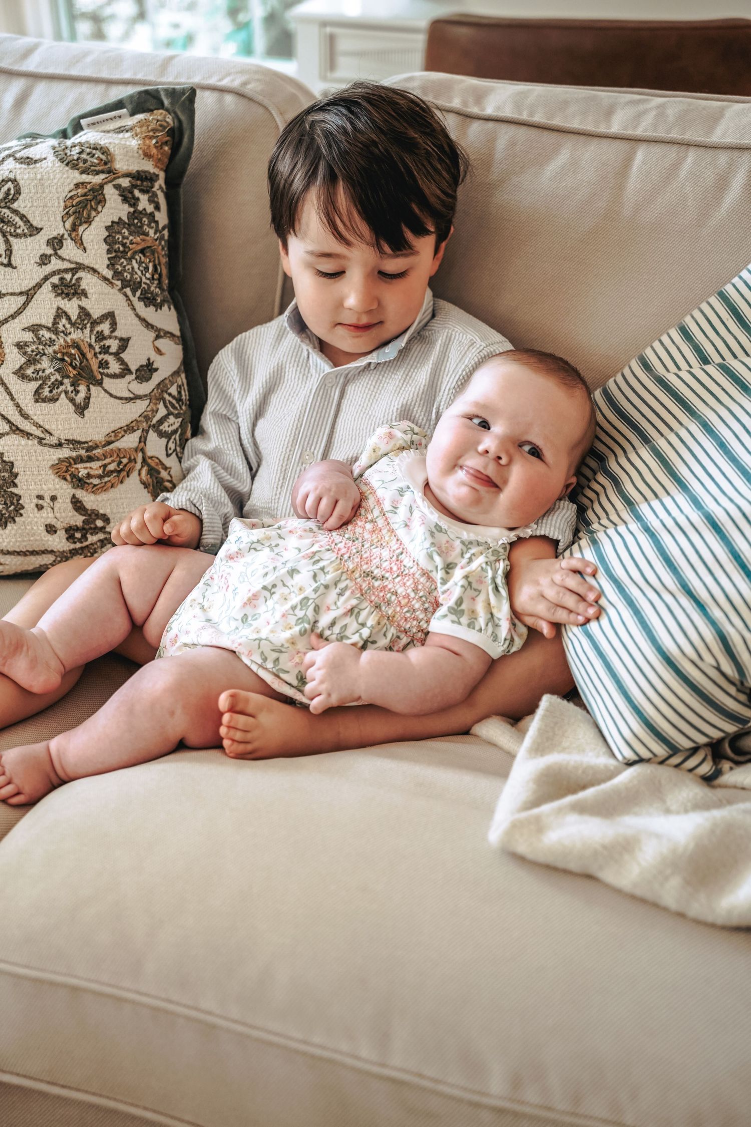 Two children sit together on a beige couch with decorative throw pillows, sharing a sweet sibling moment.