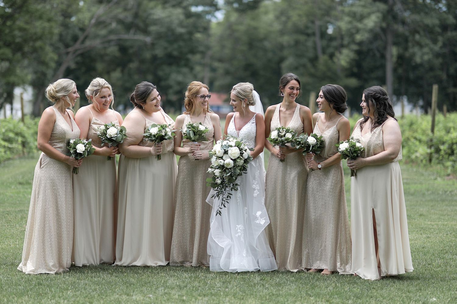 Bridal party in champagne colored dresses poses together on grassy lawn.
