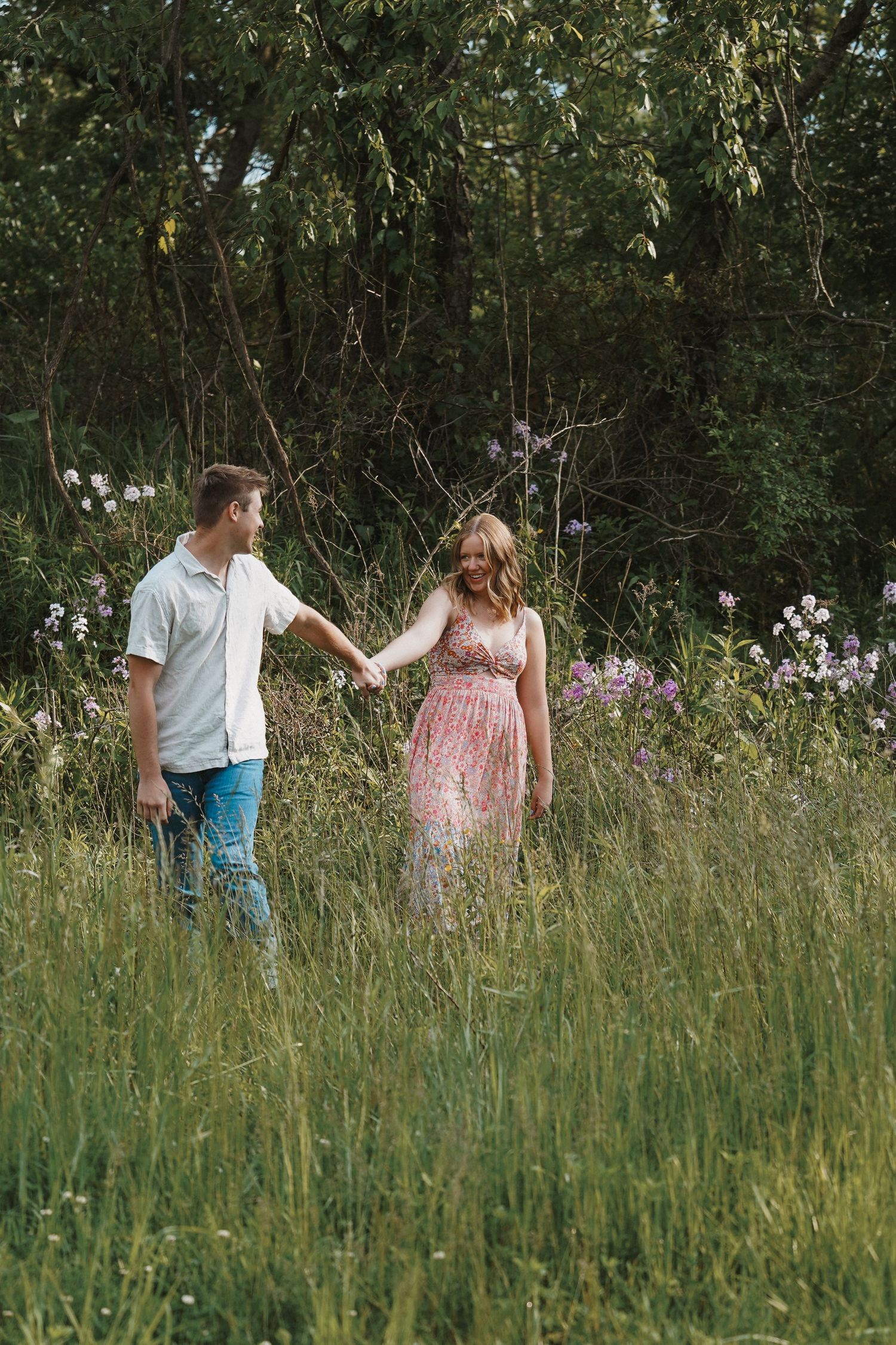 A couple holds hands while walking through a meadow with wildflowers and tall grass on a sunny day.