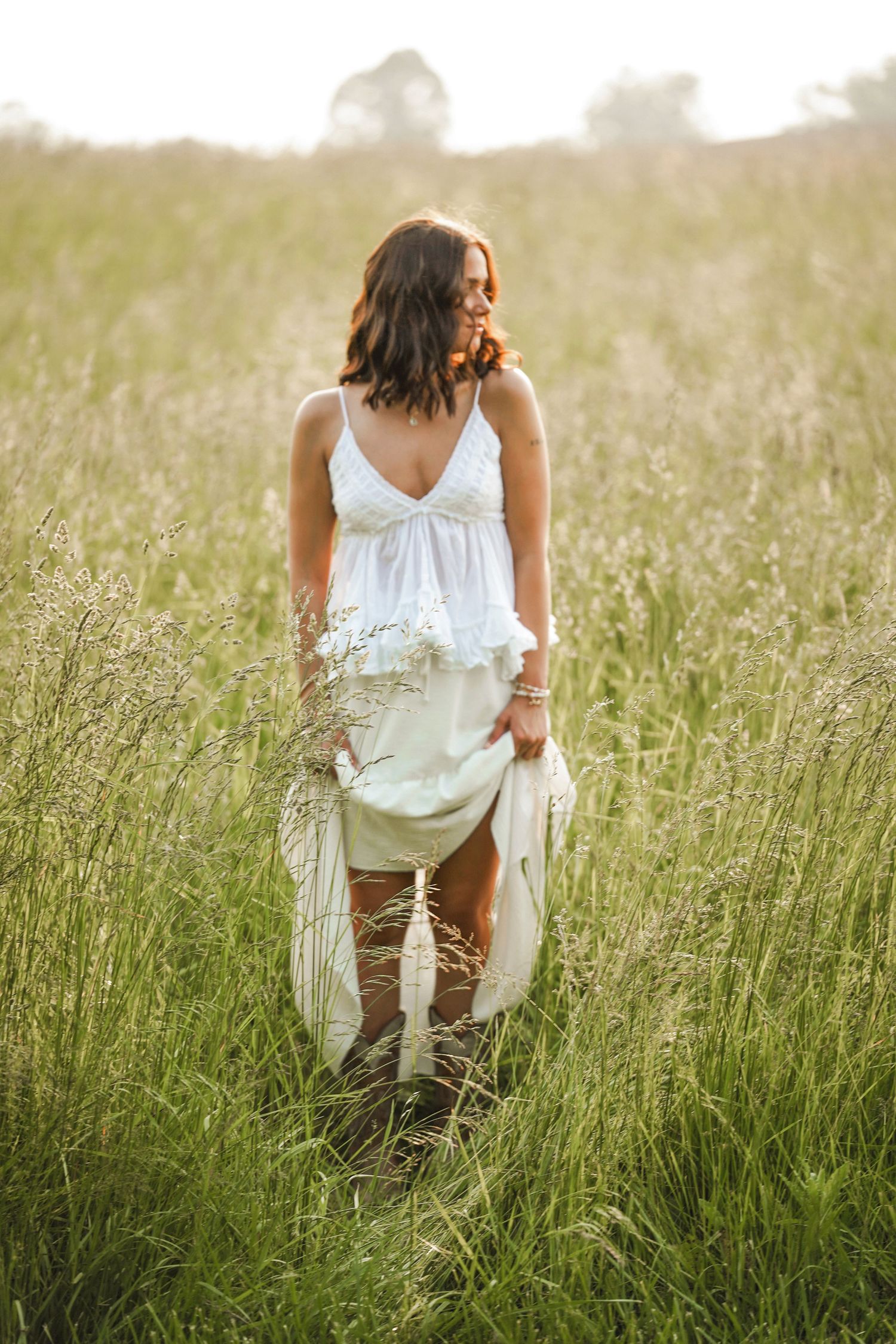 A figure in a flowing white dress stands in a sunlit meadow of tall grass during golden hour.