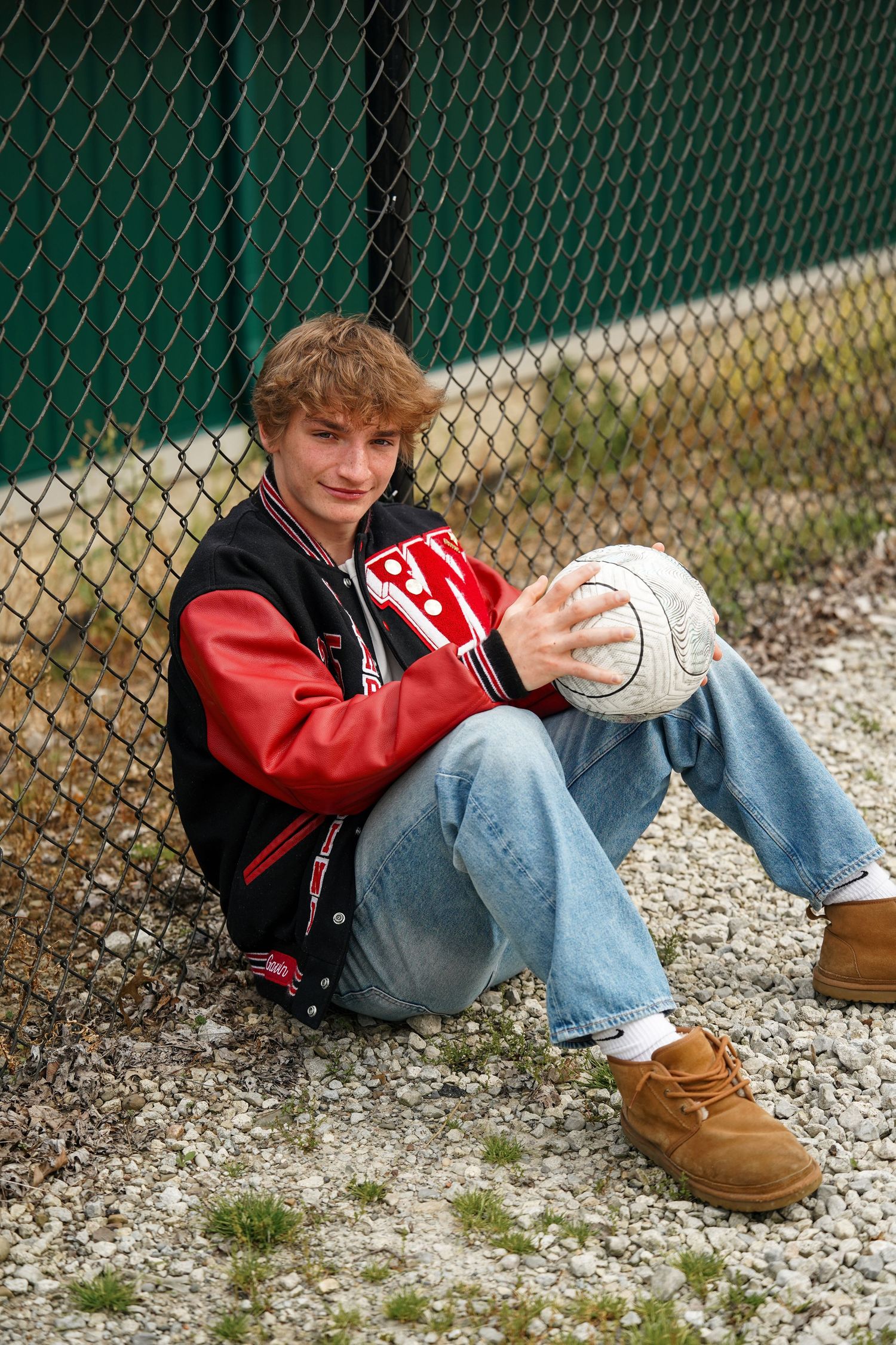 Student in varsity jacket and jeans sitting against chain link fence outdoors.