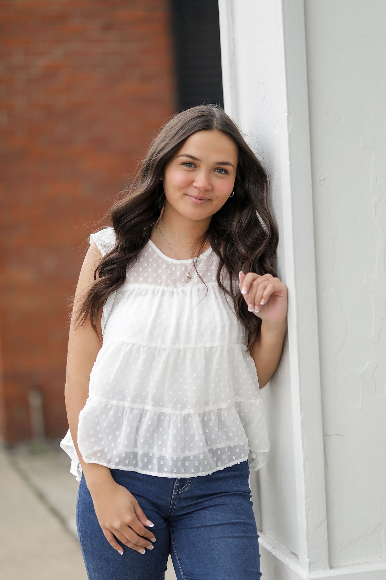 Young person in white lace top and blue jeans poses by white pillar against brick wall in casual portrait series.