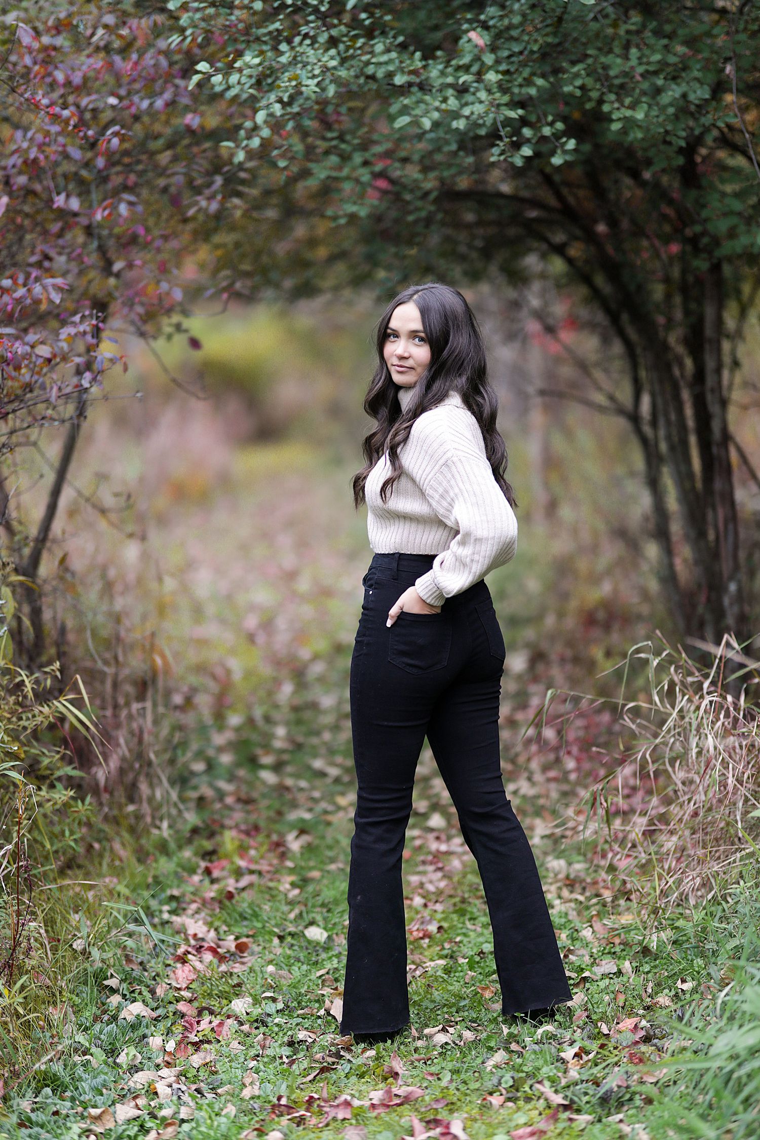 Someone wearing a white sweater and black pants poses against an autumn backdrop with blurred foliage in the background.