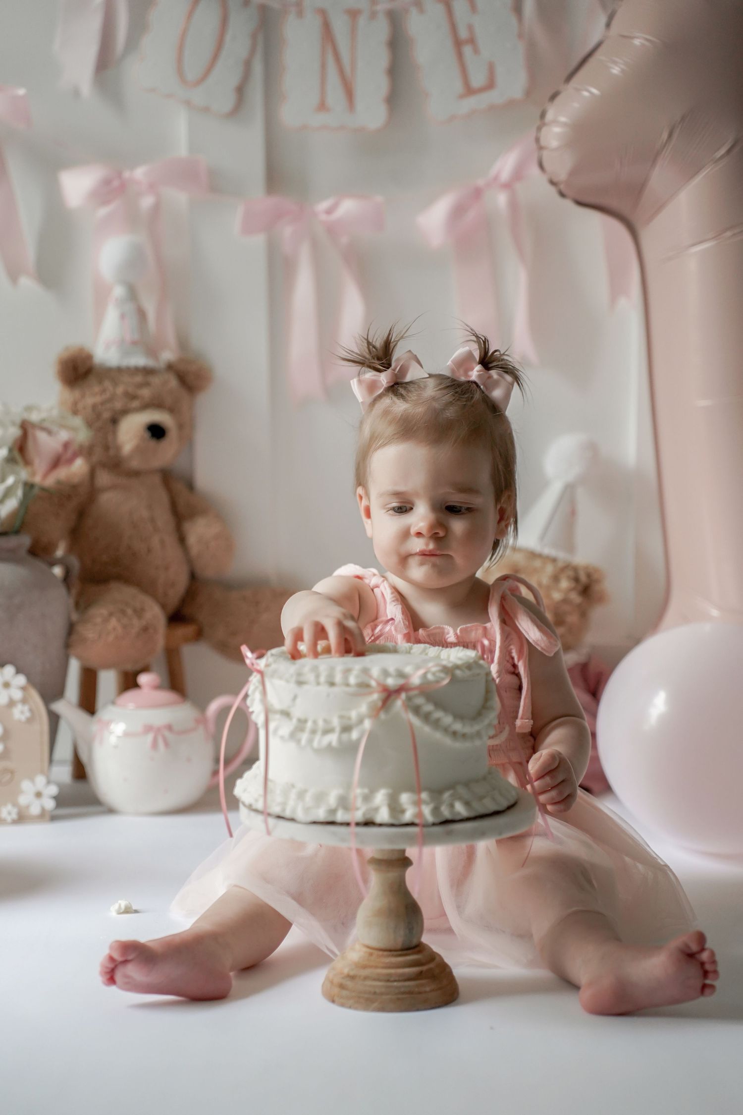 First birthday cake smash photoshoot setup in soft pink tones with white cake and teddy bear decorations.