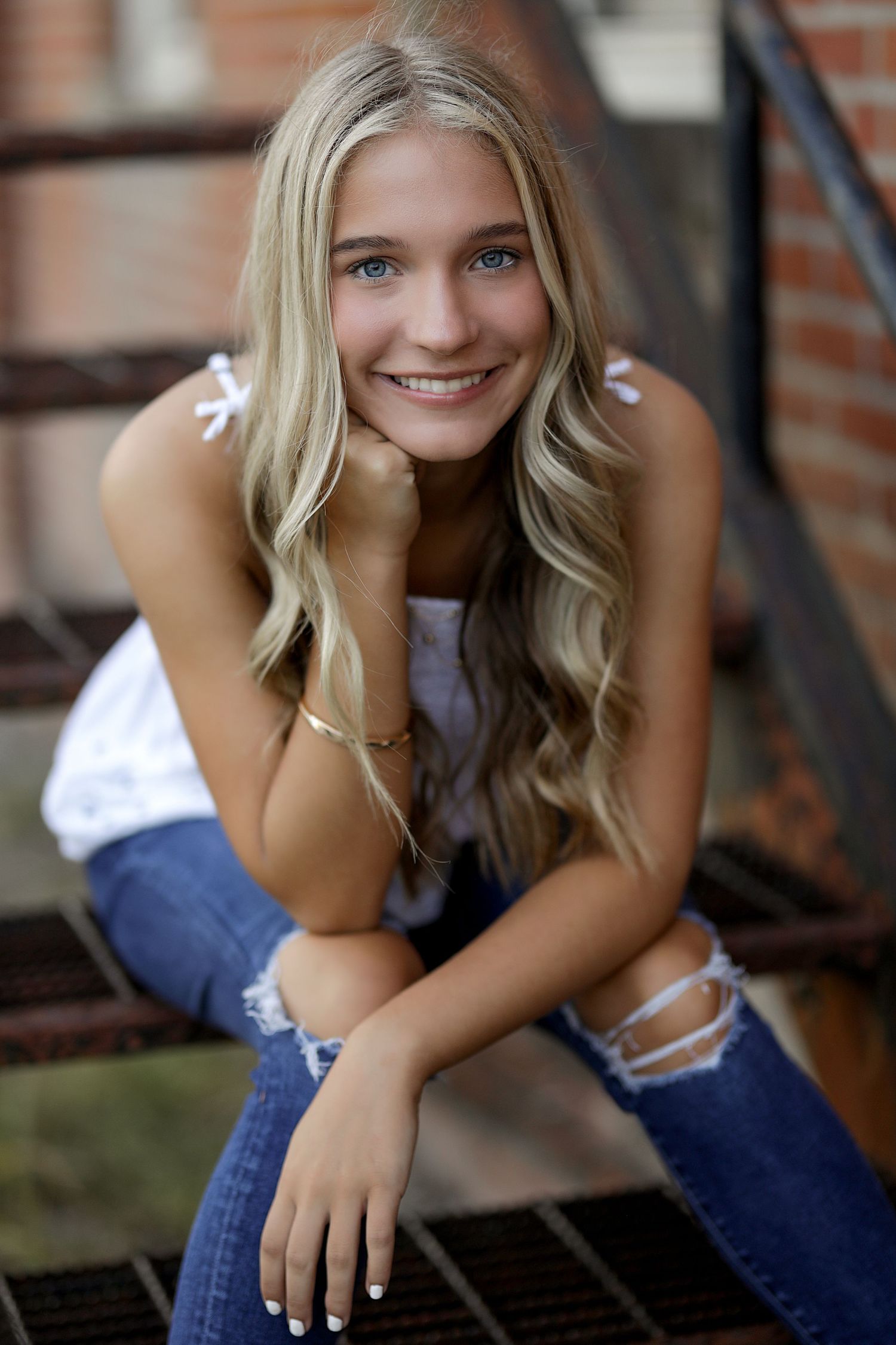 Portrait shot of a young woman in ripped jeans and white top sitting on steps by a brick wall, smiling warmly at camera.