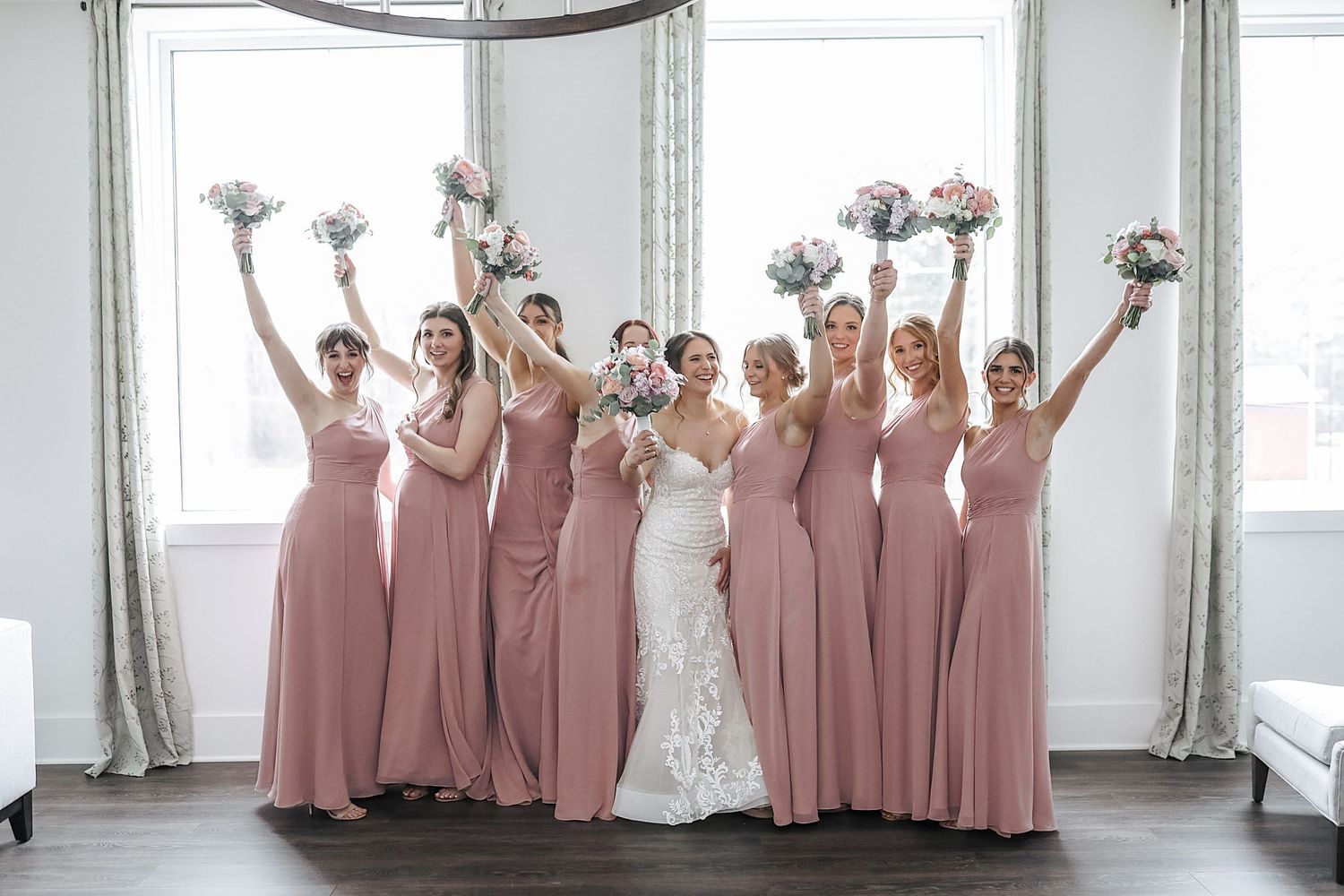 A wedding party in dusty rose bridesmaid dresses poses together in a bright white room with windows.