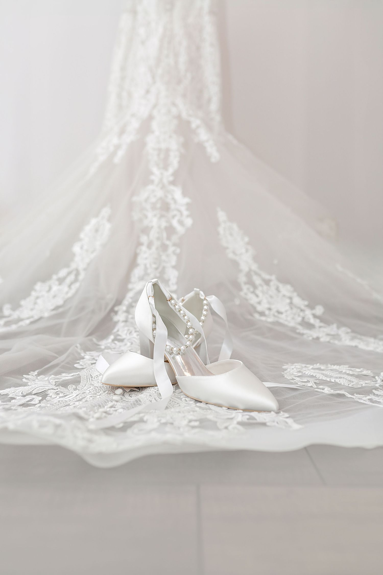 Elegant white bridal shoes displayed on delicate lace veil with intricate floral embroidery details.