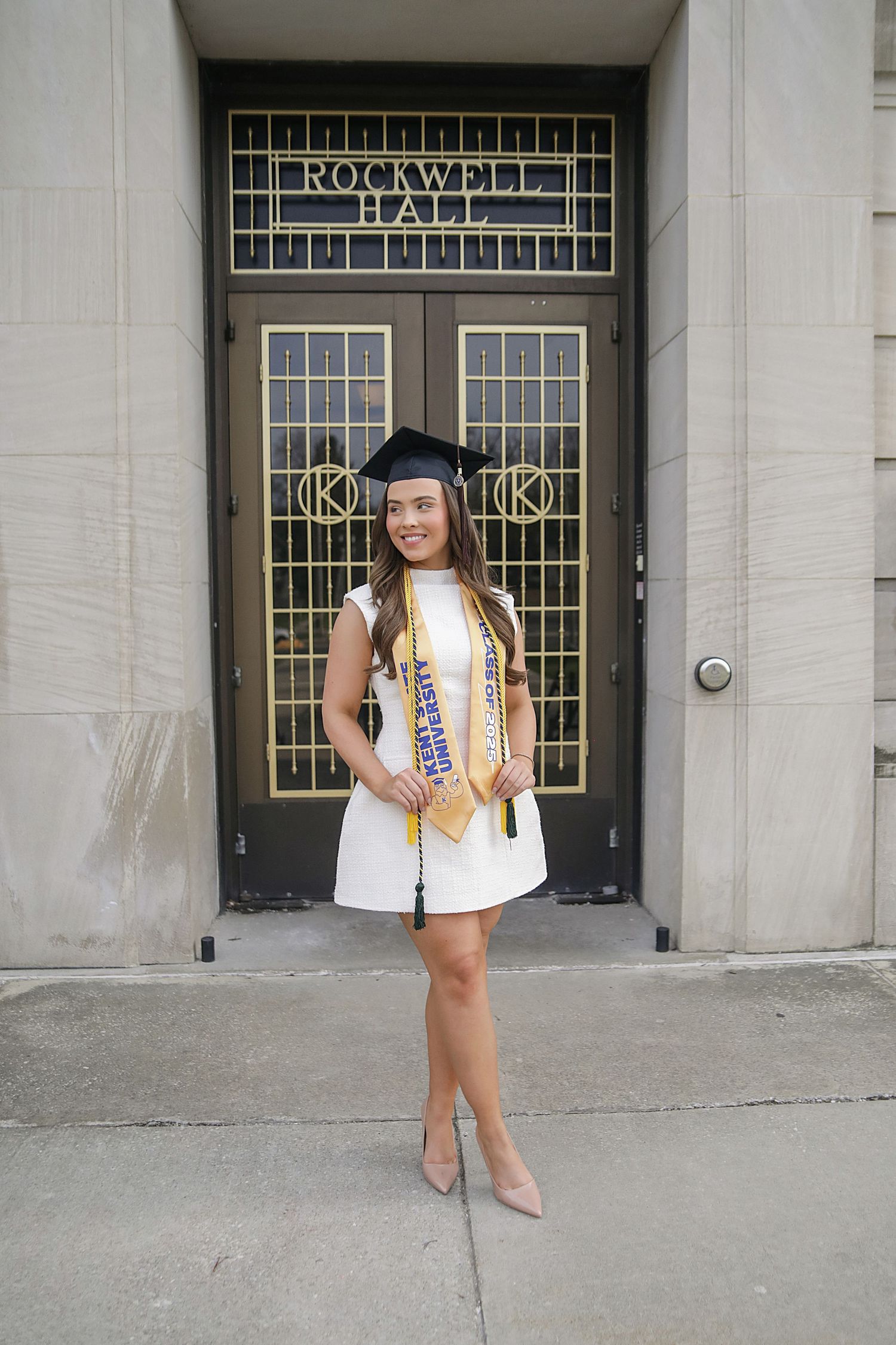 Graduate in cap and gown poses in front of Rockwell Hall entrance wearing gold stole and white top.