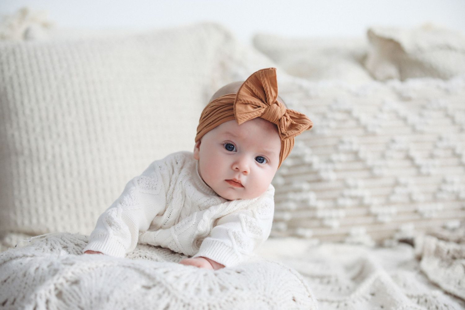 A baby in white clothing lying on a textured white blanket with a bow hair accessory.