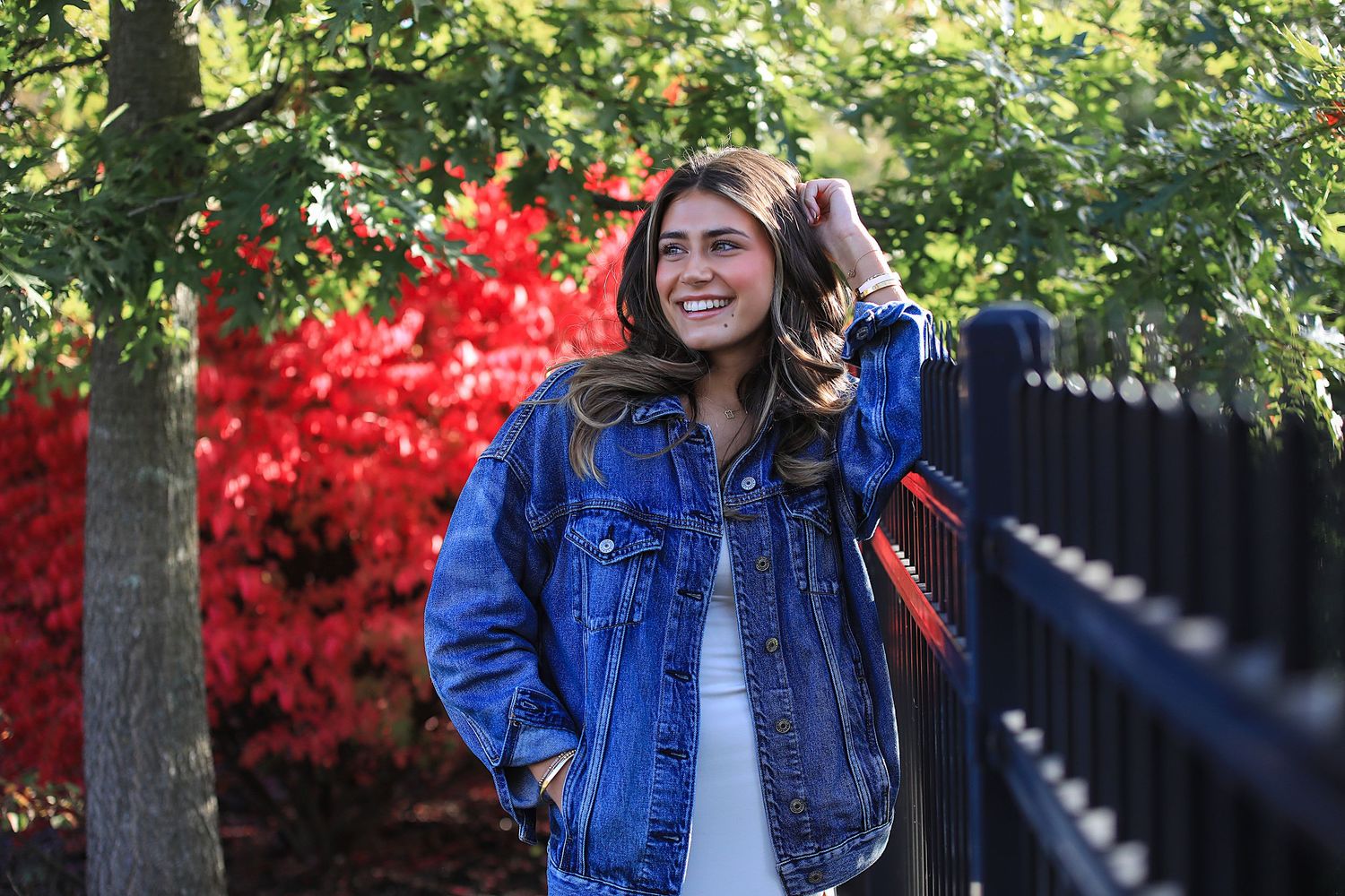 A figure in a denim jacket poses joyfully in front of vibrant fall foliage and autumn trees.
