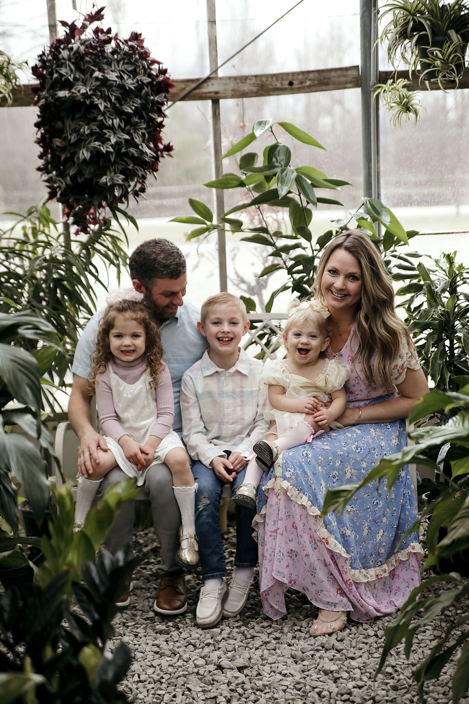 A joyful group sits together and laughs while spending time in a greenhouse filled with lush plants and greenery.