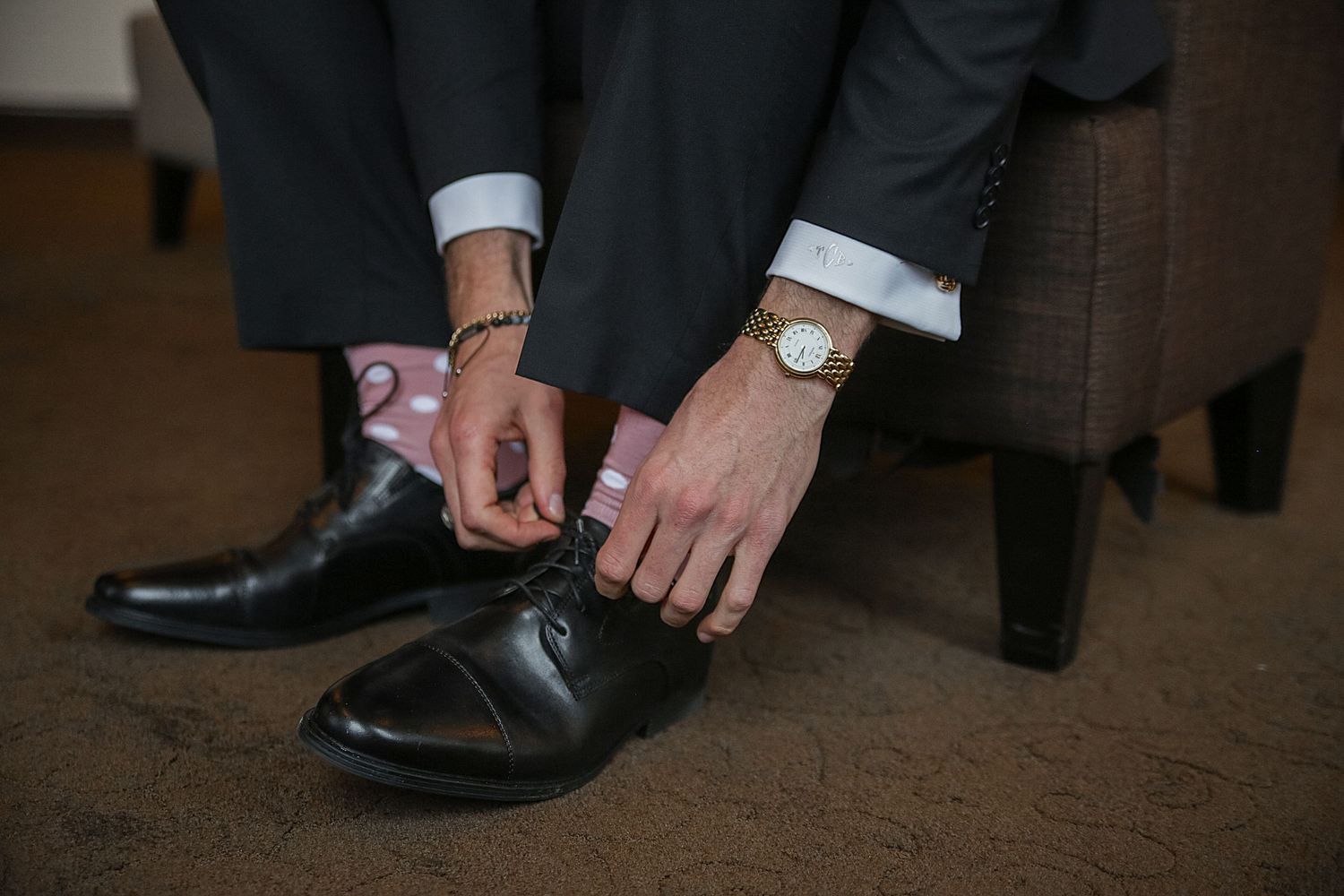 Close-up view of hands tying black leather dress shoes while wearing a dark suit with white watch and cuffs.