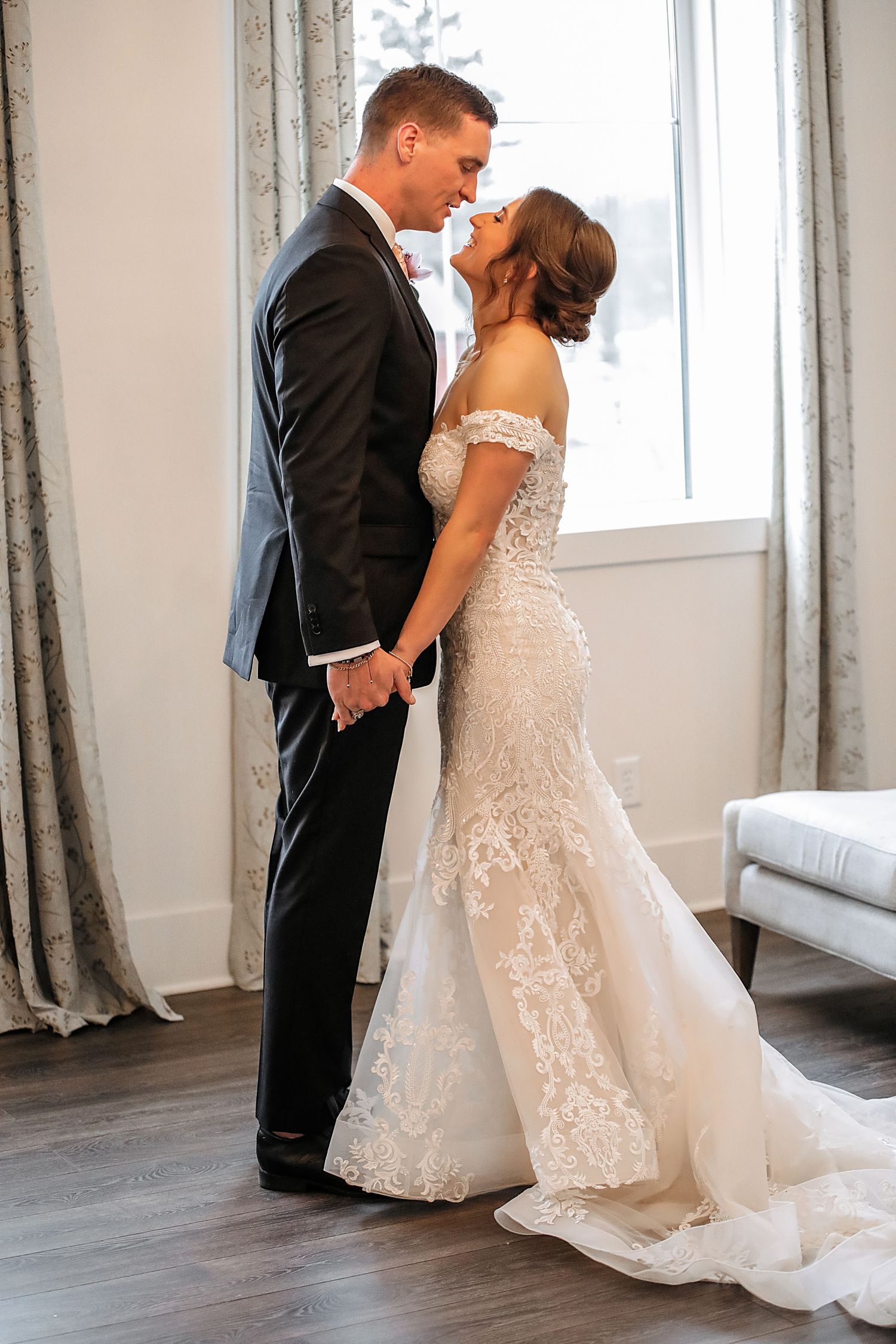 Romantic wedding couple sharing intimate moments in elegant white room with floor-to-ceiling windows and curtains.