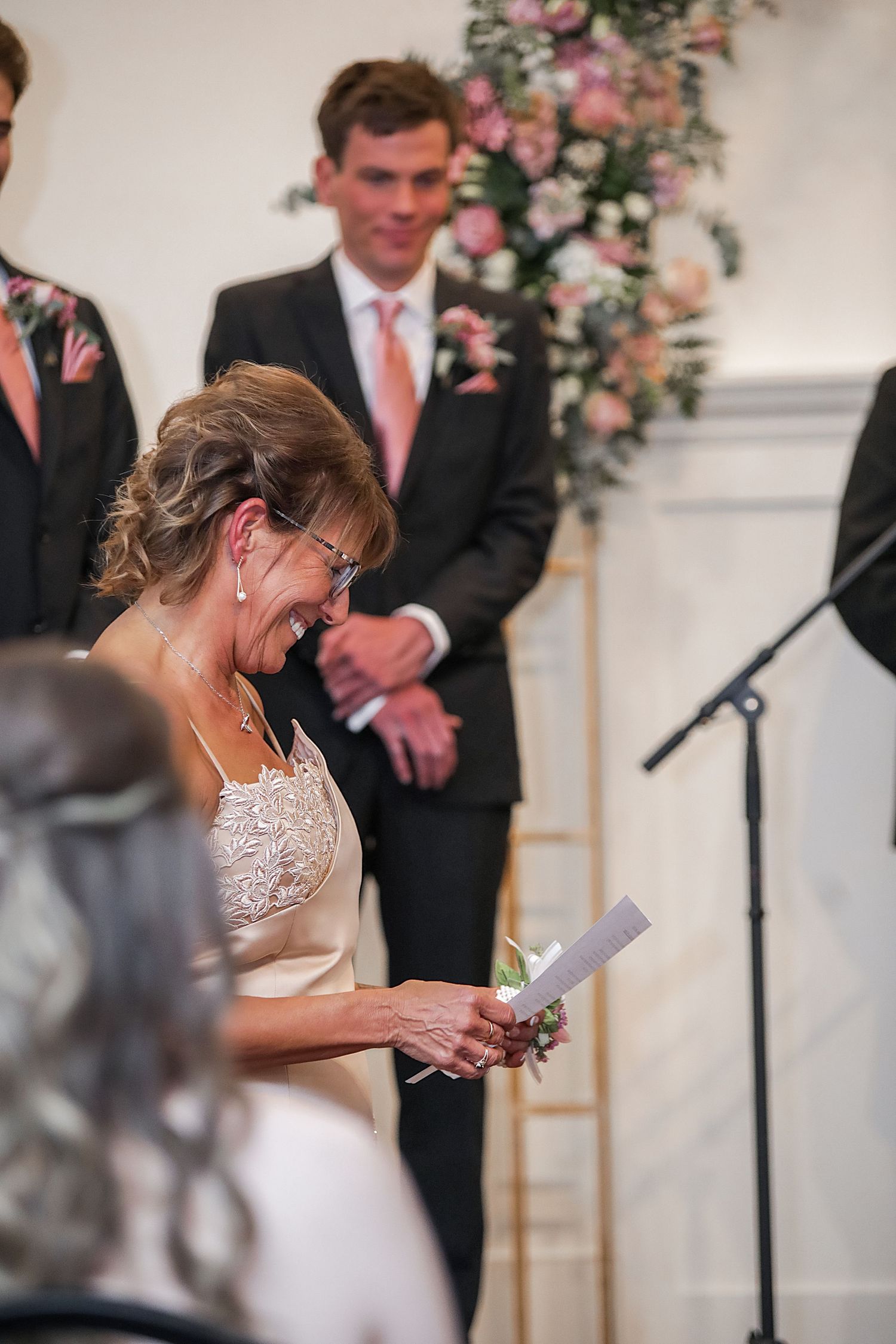 A bride in a lace wedding dress reads from papers during a ceremony with pink floral decorations in the background.