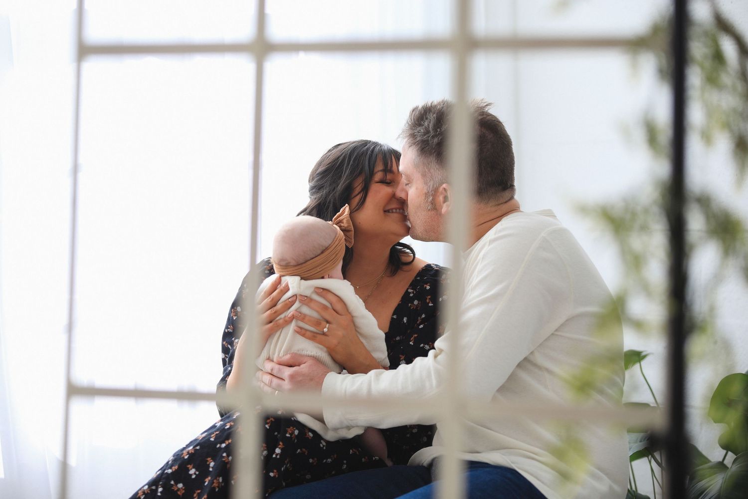 A couple in formal wedding attire shares an intimate kiss by a window with natural light streaming in.
