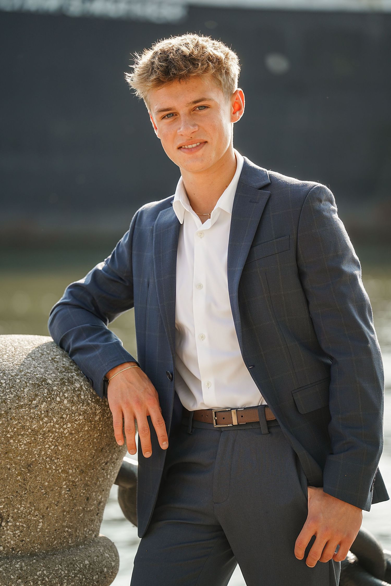 Professional portrait of someone in a navy suit and white dress shirt leaning against a stone railing outdoors.