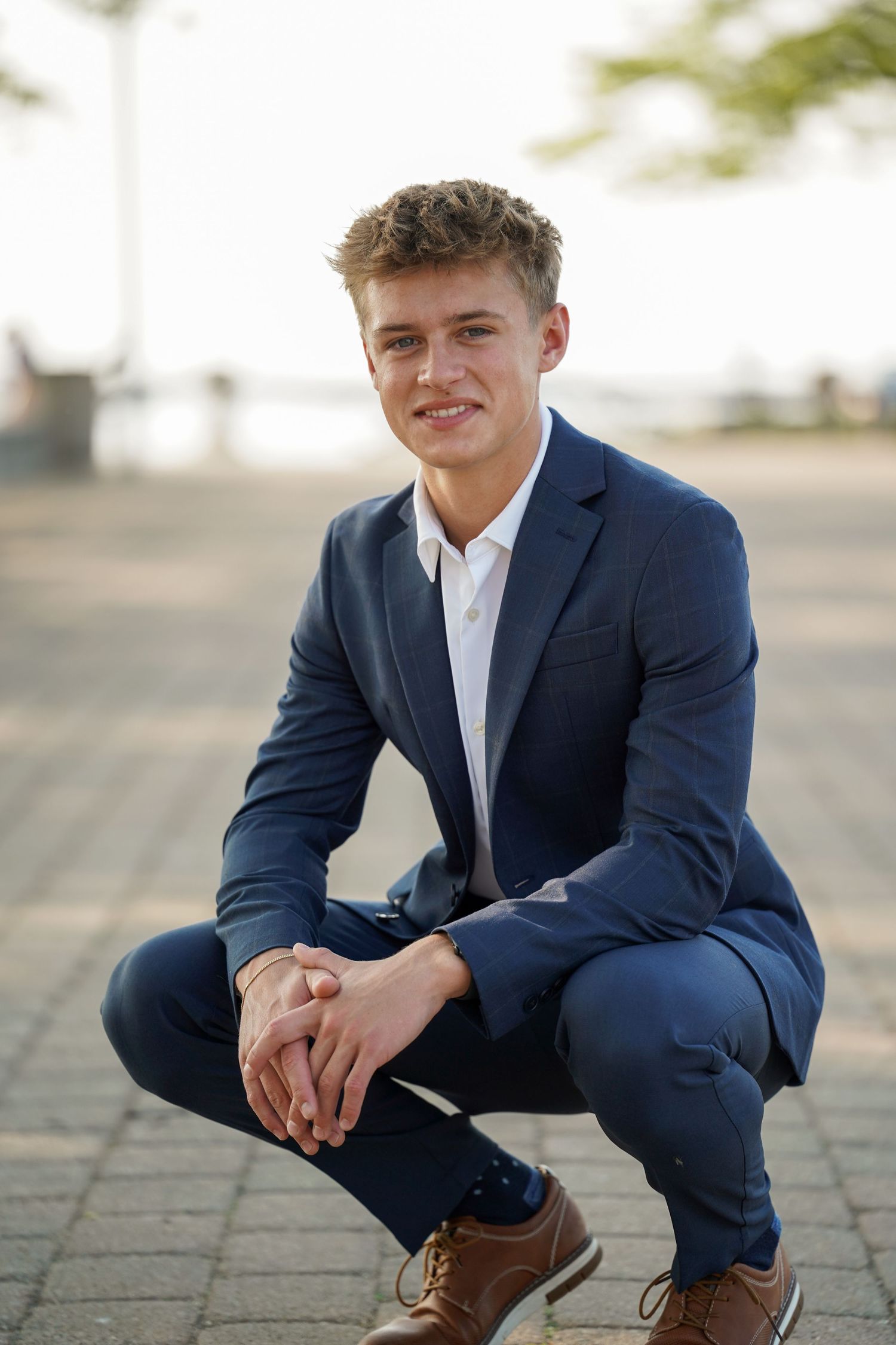 Young professional in dark blue suit and brown dress shoes crouches down while posing outdoors for a portrait.