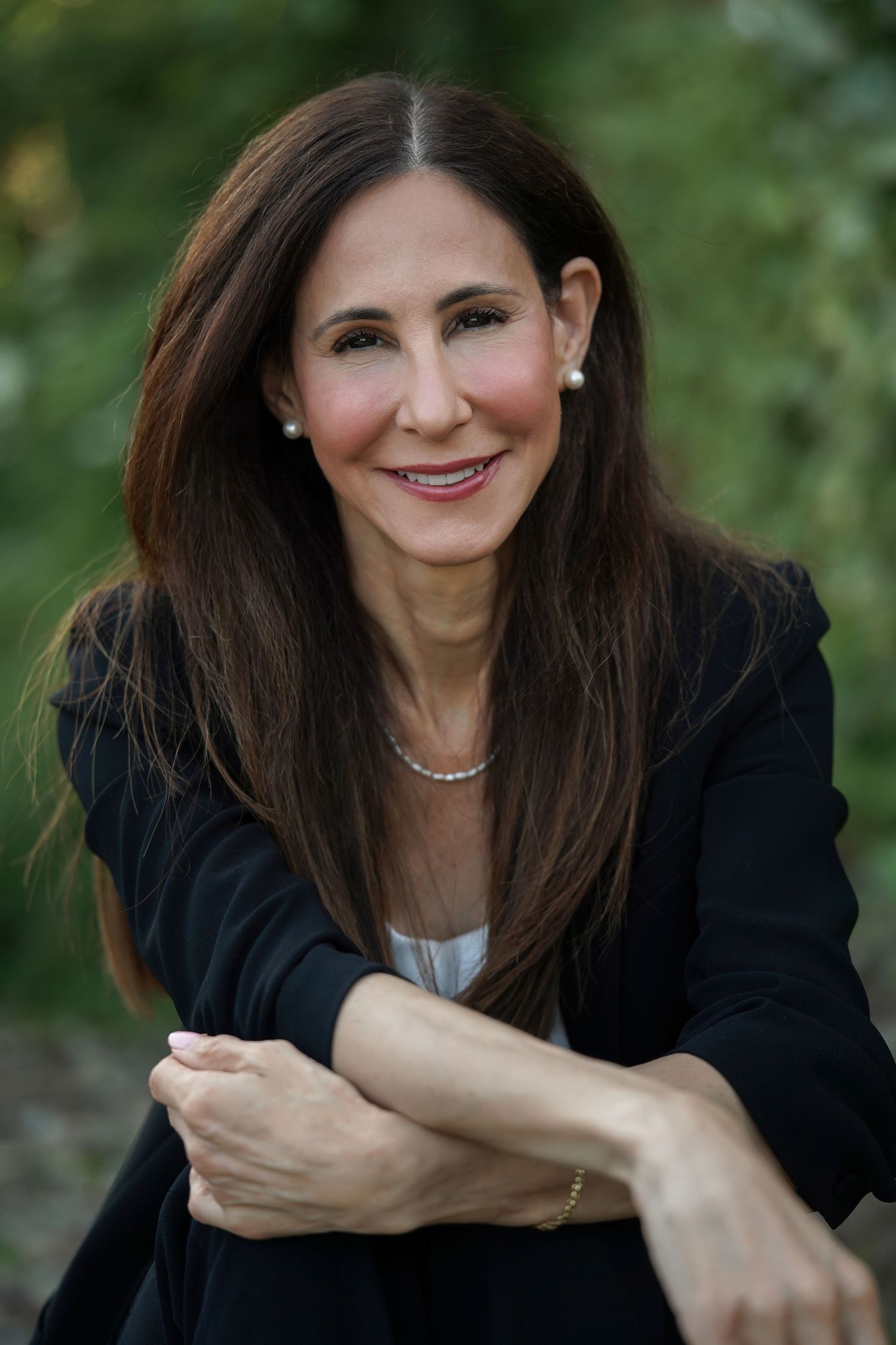 Professional portrait of a woman with long dark hair wearing a black blazer and pearl necklace smiling outdoors.