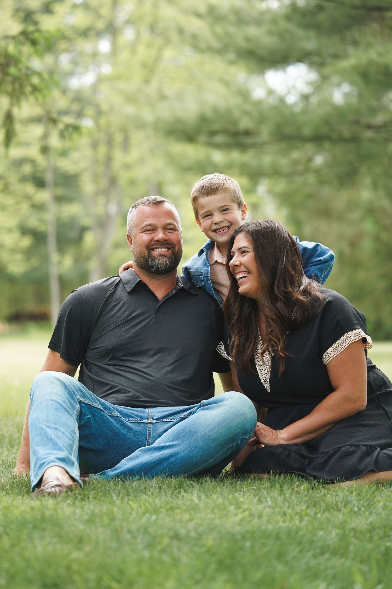 A happy family shares a joyful moment while sitting together on green grass in a park-like setting with trees in background.