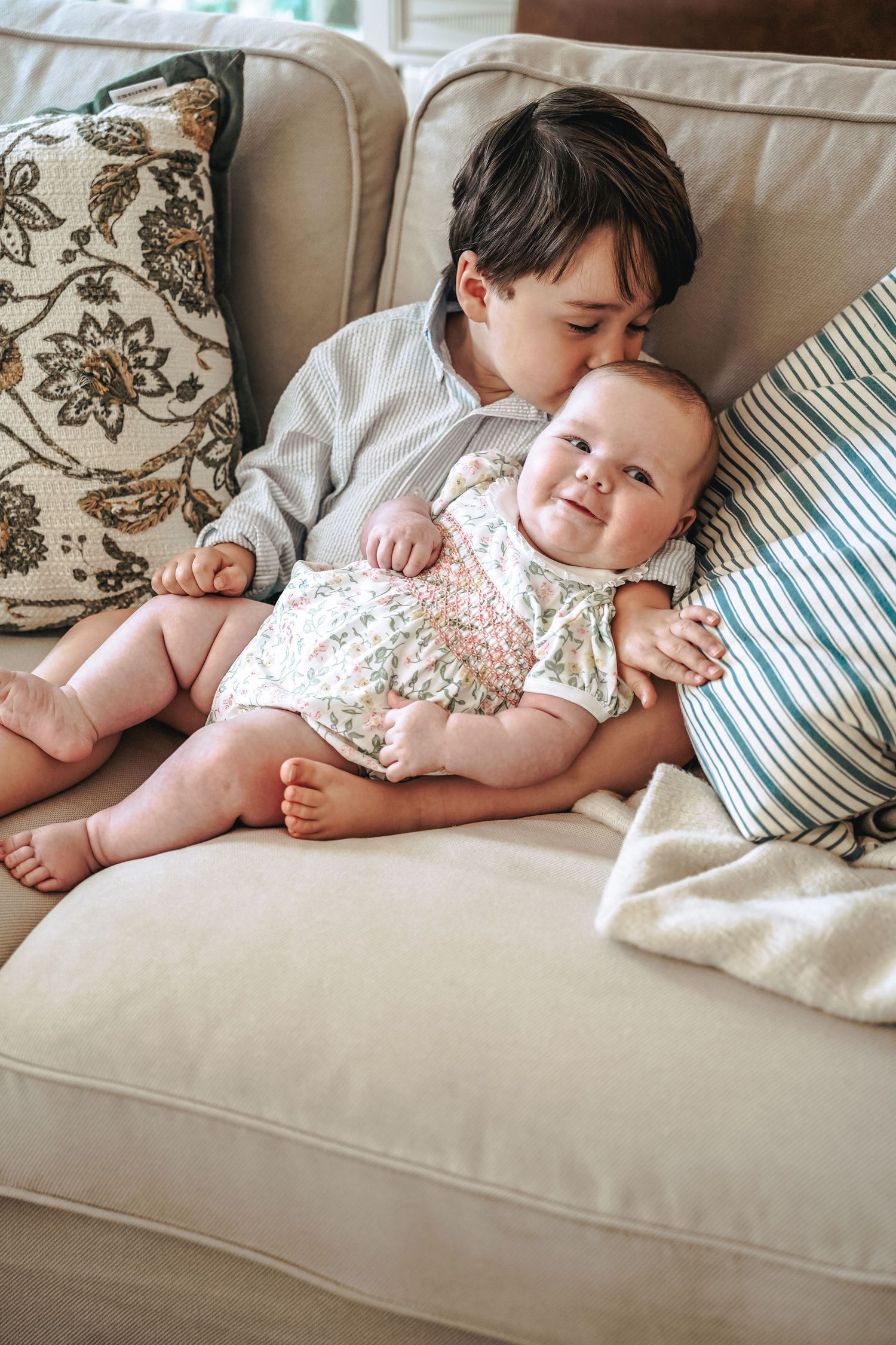Two children sit together on a beige couch with floral pillows, sharing a sweet moment of bonding.